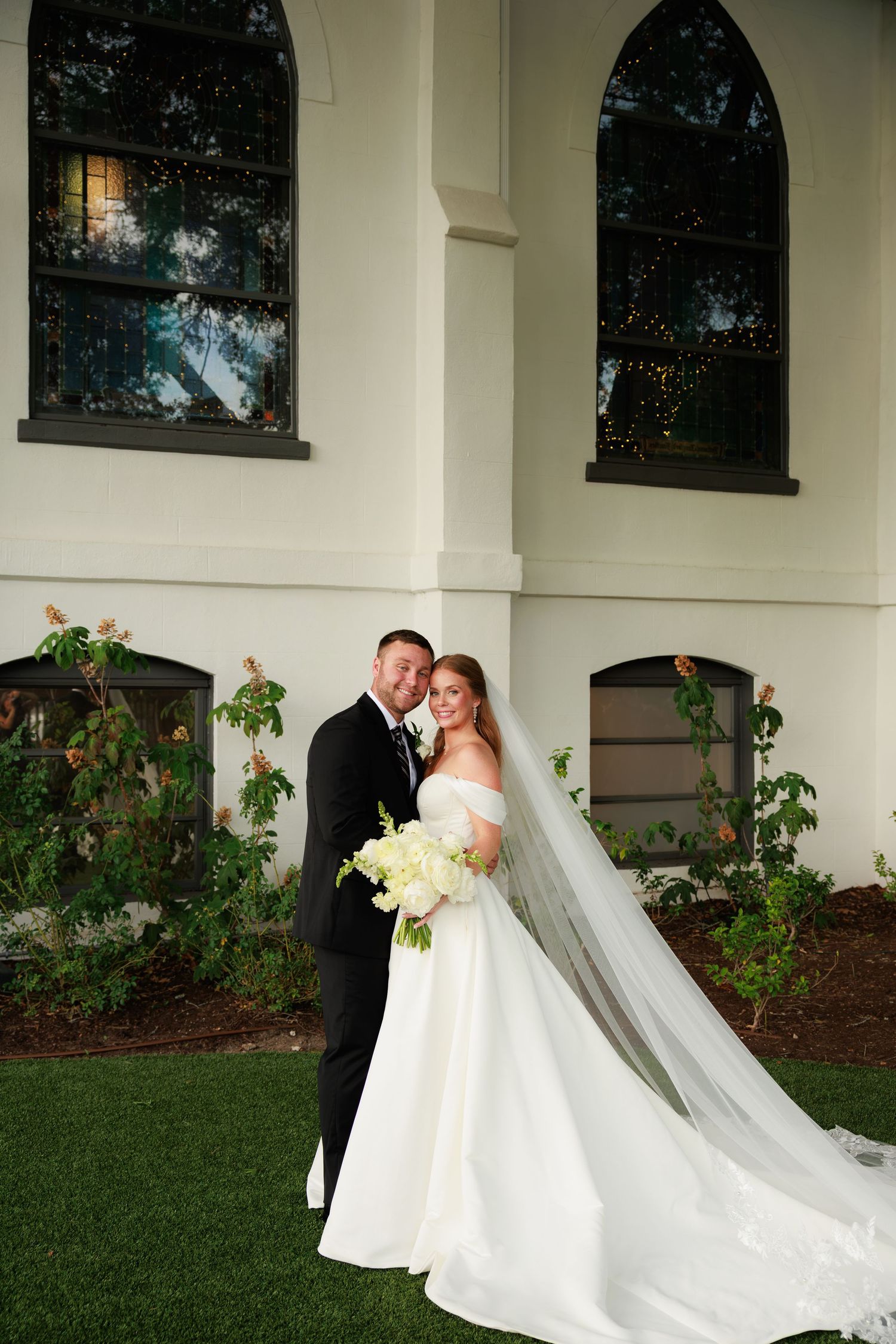 Bride and groom portrait outside white chapel, bride in off-shoulder gown with long veil holding white bouquet.