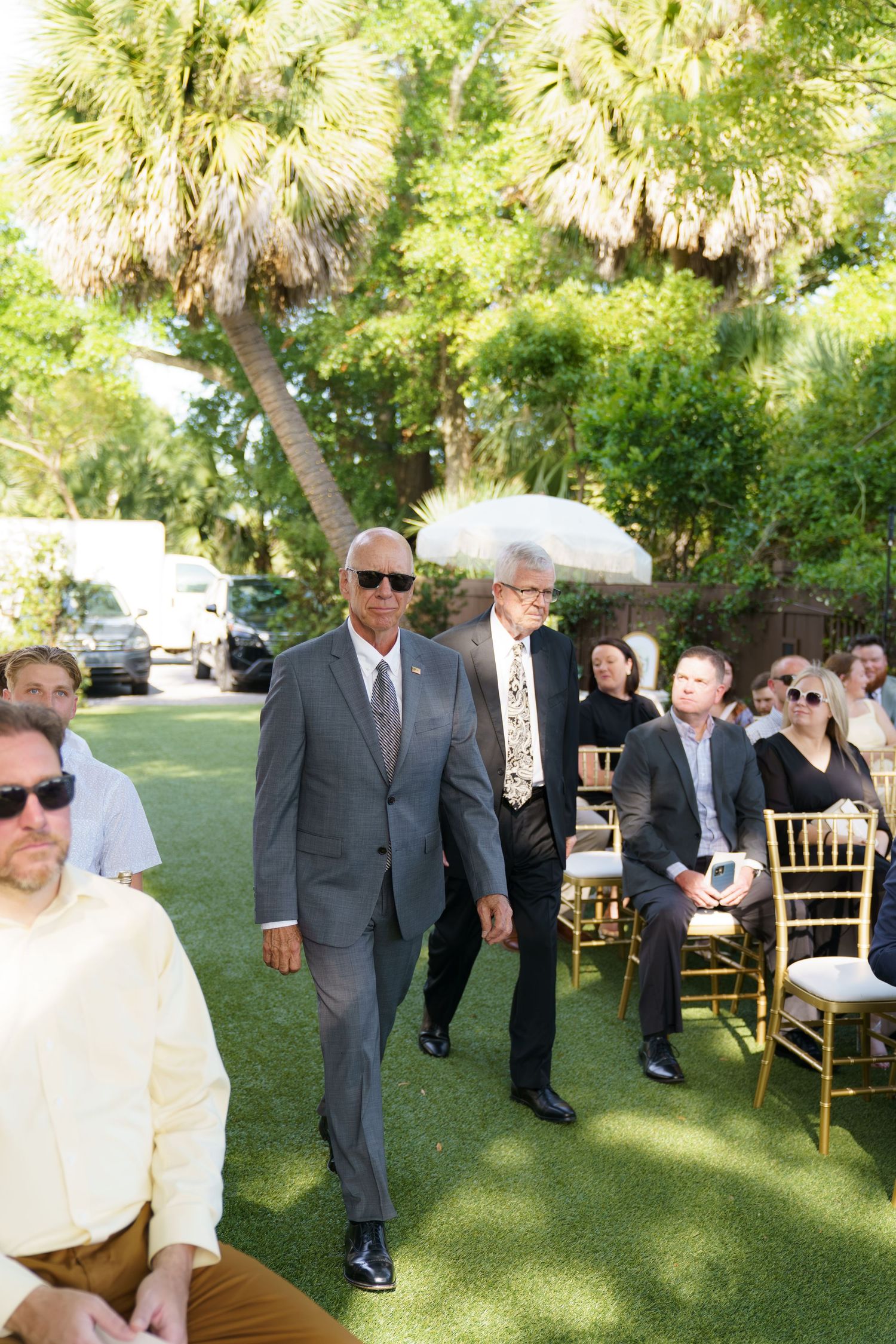 Two men in gray suits walk down outdoor wedding ceremony aisle.