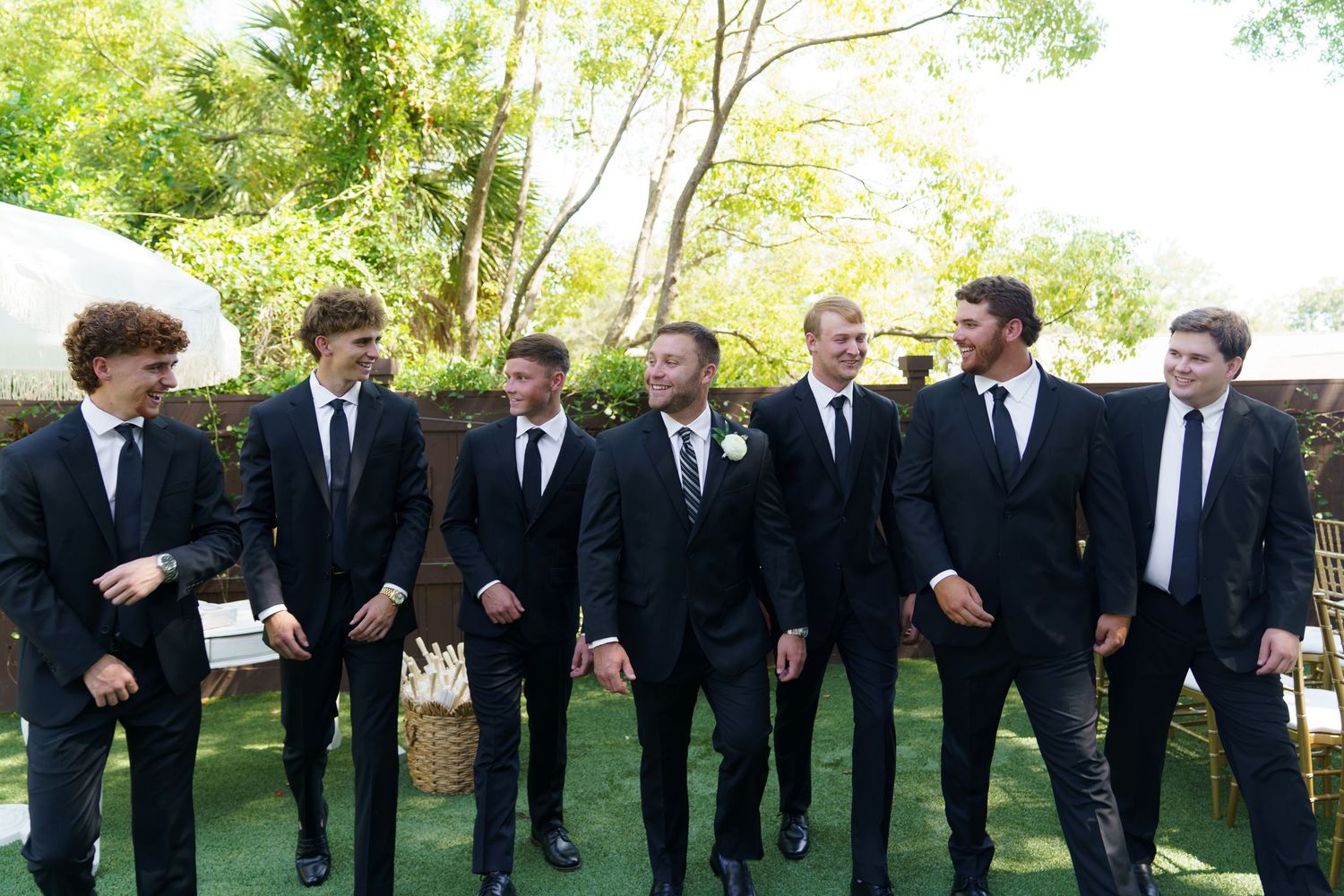 Seven groomsmen in black suits laughing and walking together outdoors at a wedding celebration.