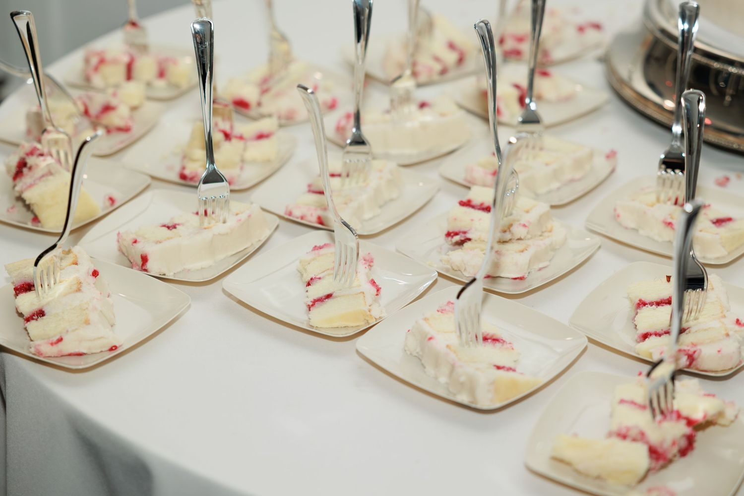 Elegant dessert table with multiple white square plates of white cake slices topped with red berry sauce and forks.