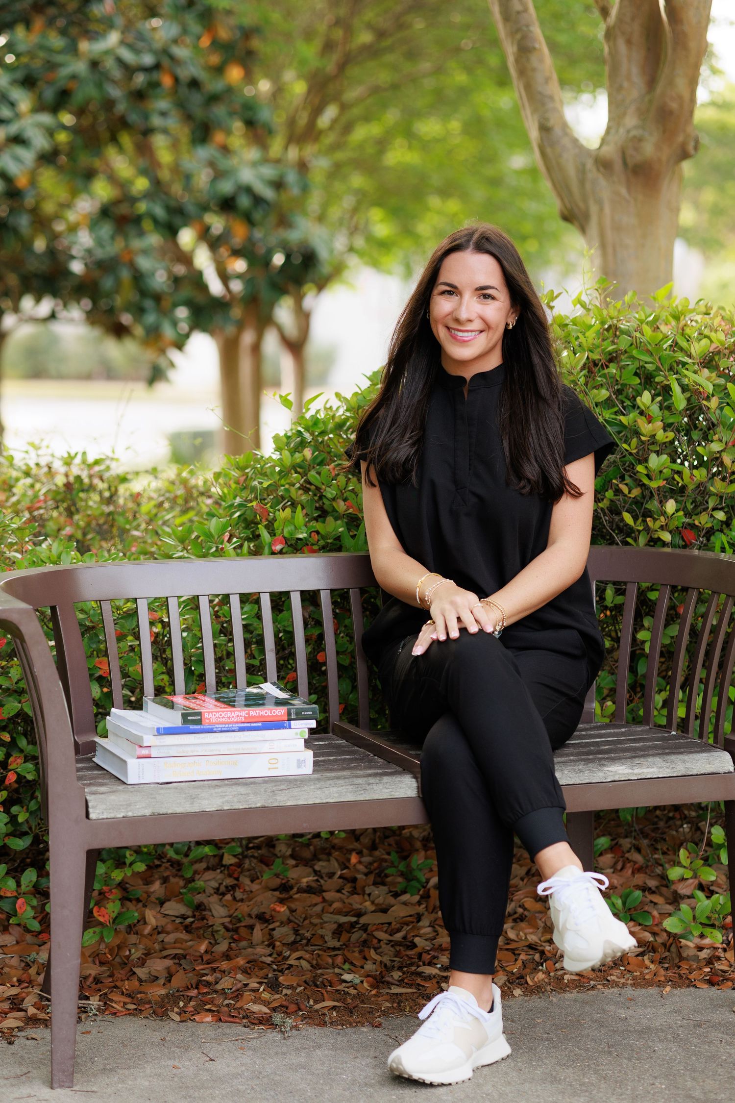 Woman in black outfit sitting on a bench outdoors with stacked books, smiling in a lush green garden setting.