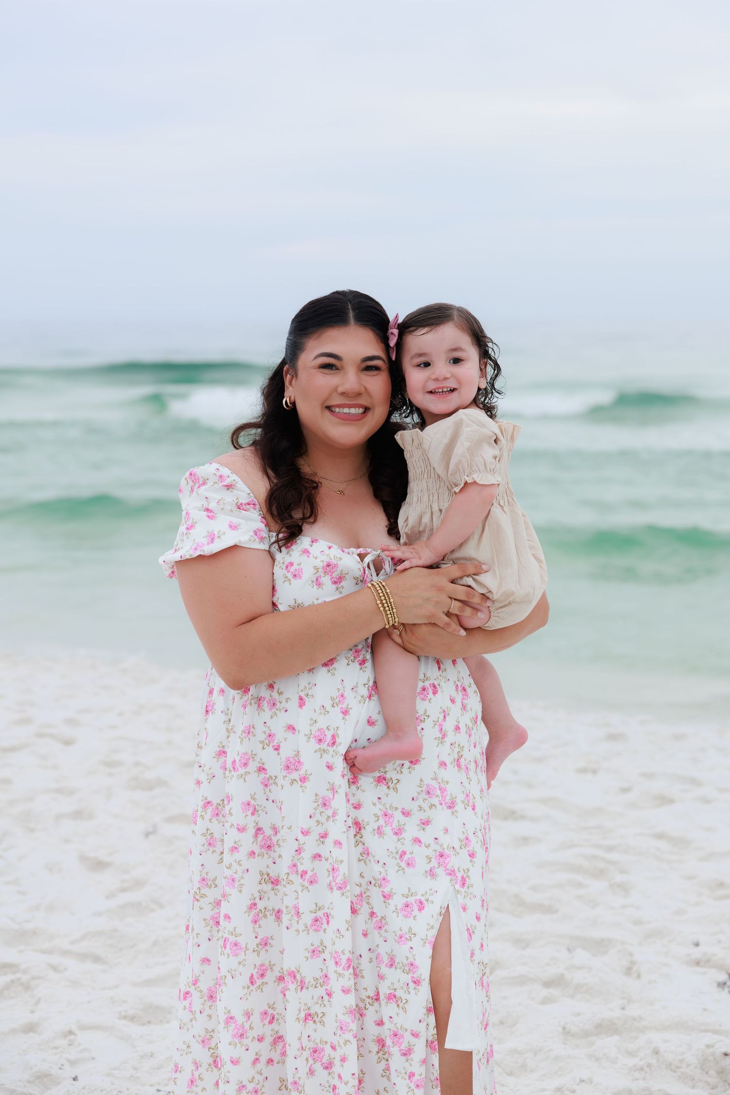 A smiling woman in a floral dress holds a baby girl on a white sand beach with turquoise waves.