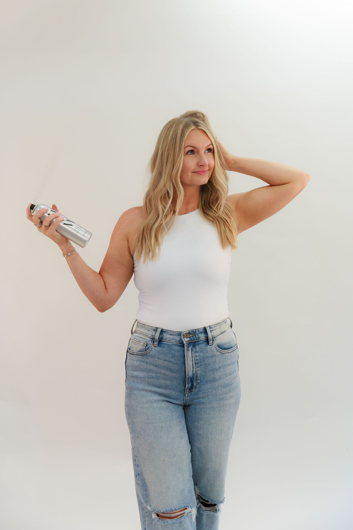 Woman in white tank top and ripped jeans holding styling product against white background.