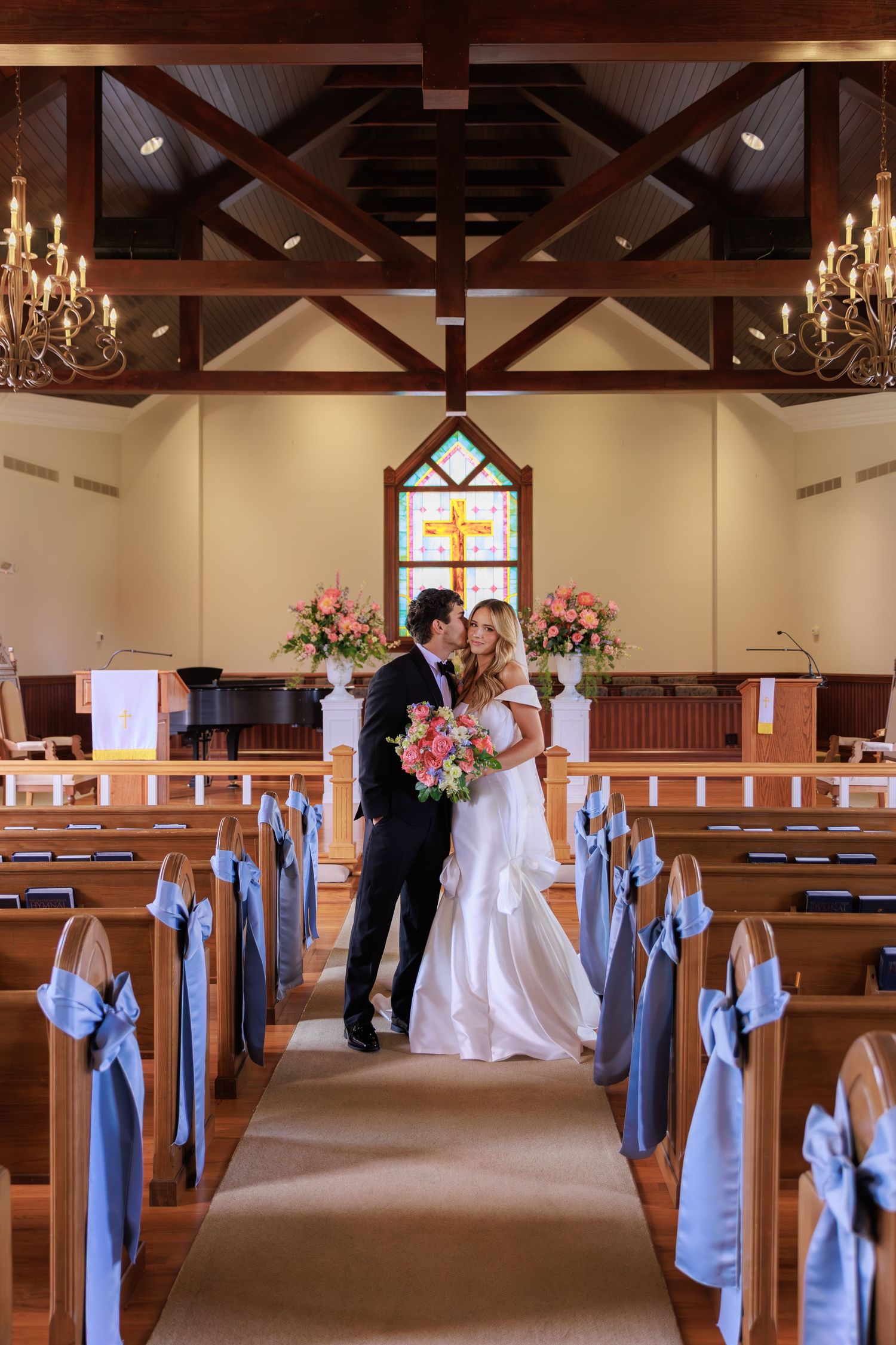 A wedding couple stands in a church aisle decorated with light blue bows, chandeliers and floral arrangements.