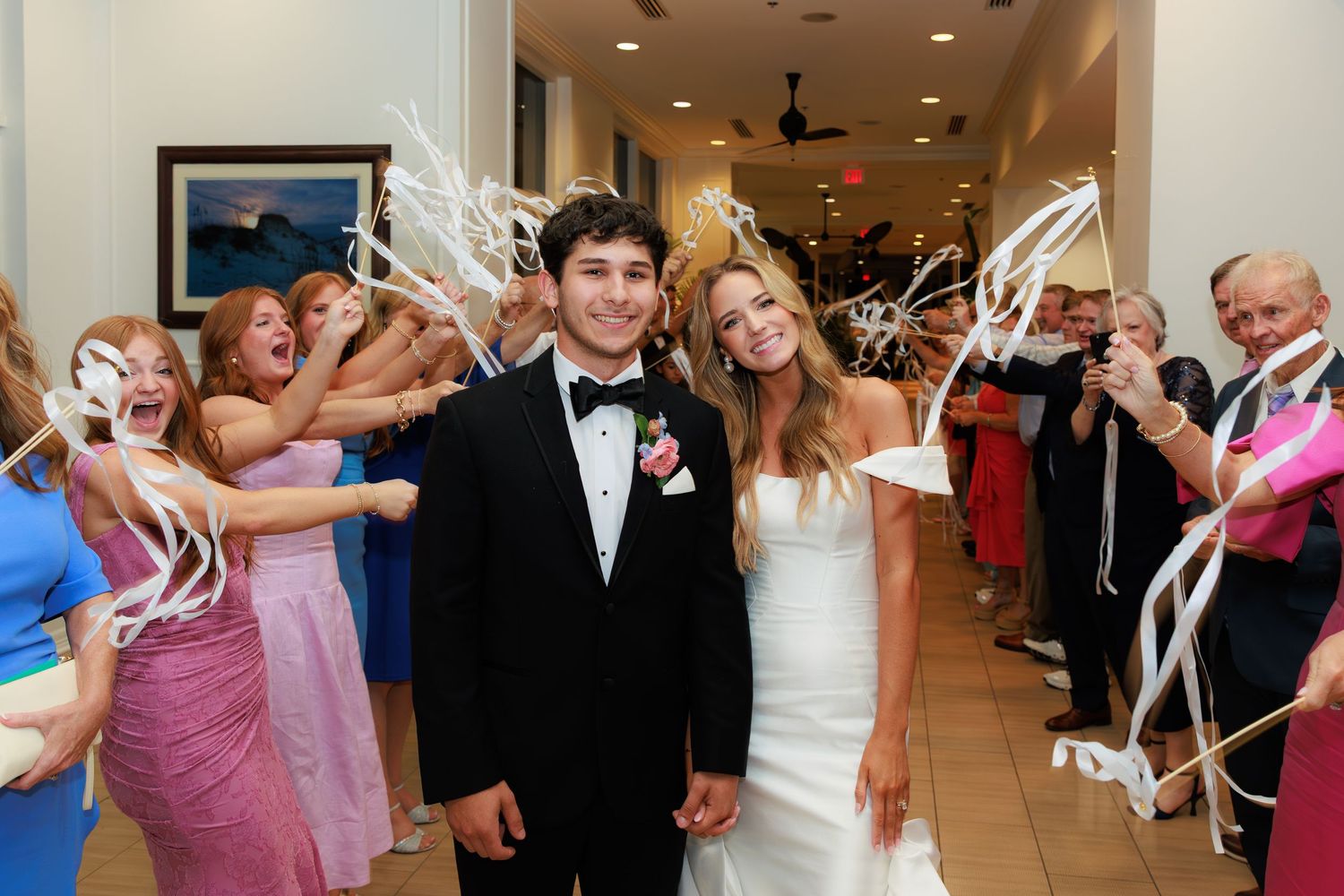 Wedding couple walks through corridor as guests wave white streamers during celebratory exit from ceremony.