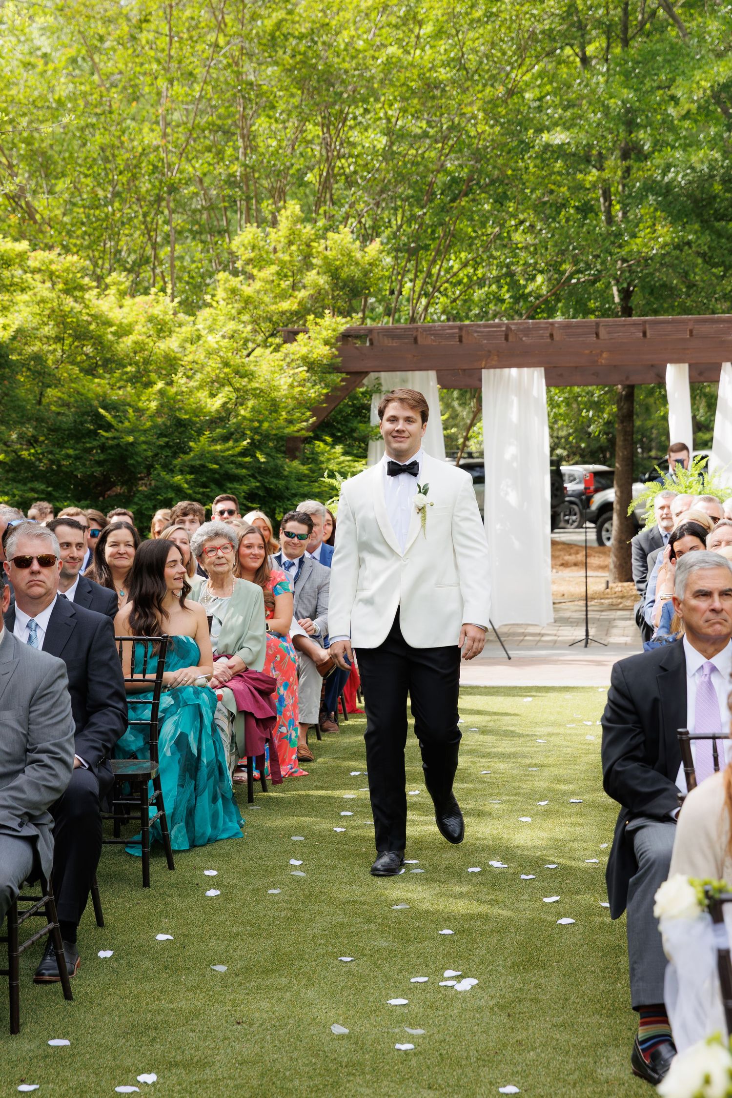 Groom in white tuxedo jacket walks down outdoor wedding ceremony aisle smiling at guests.