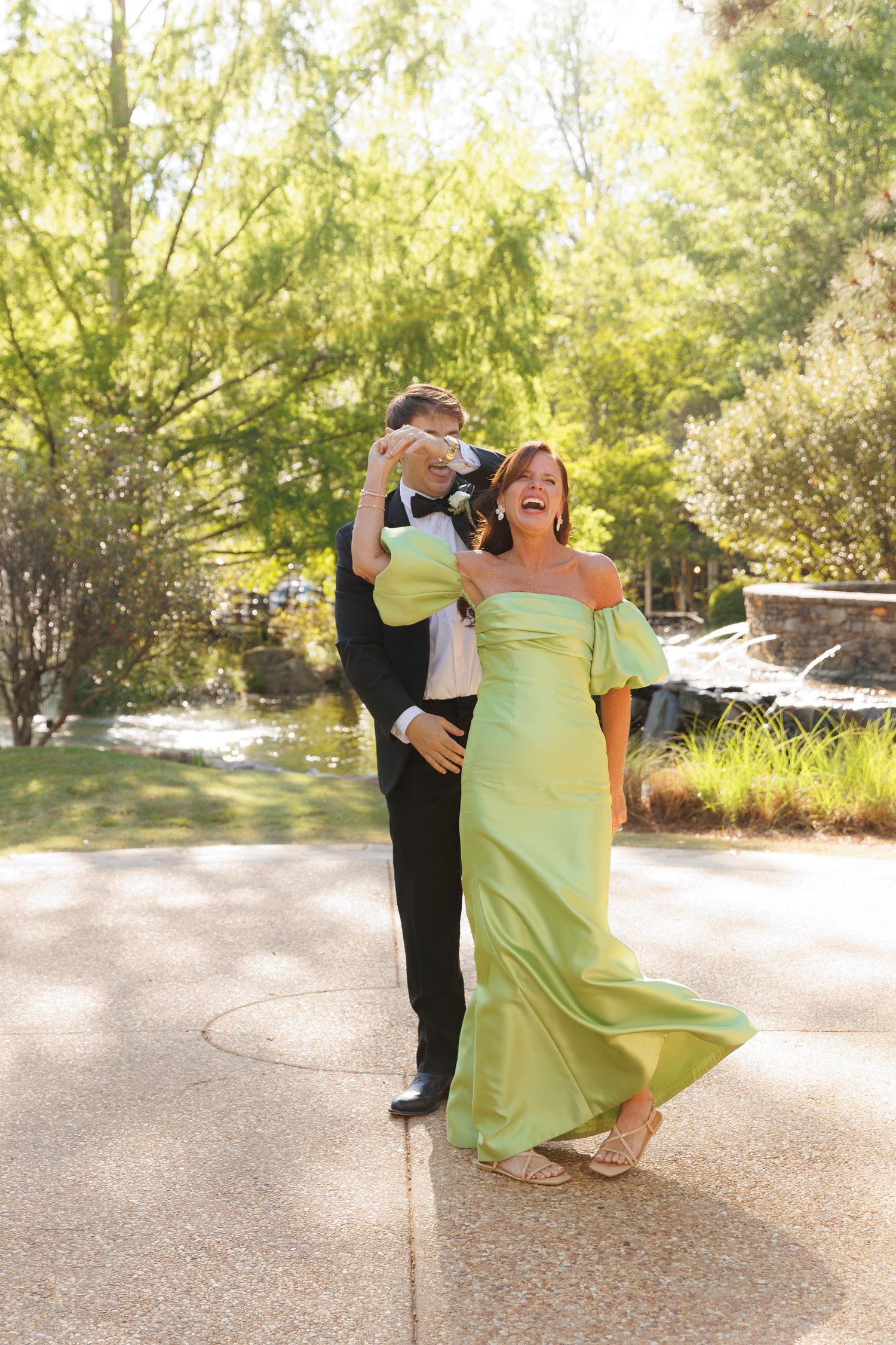 Couple laughing and dancing outdoors, woman wearing an elegant off-shoulder sage green gown at a formal event.