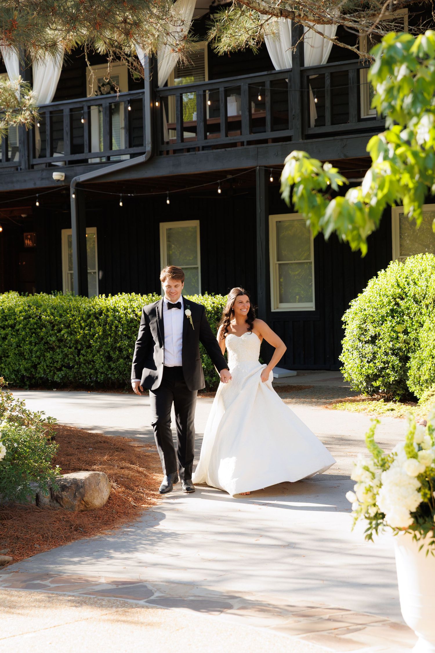 Bride in white gown and groom in black tuxedo walking hand in hand outside elegant dark-wood venue with lush greenery.