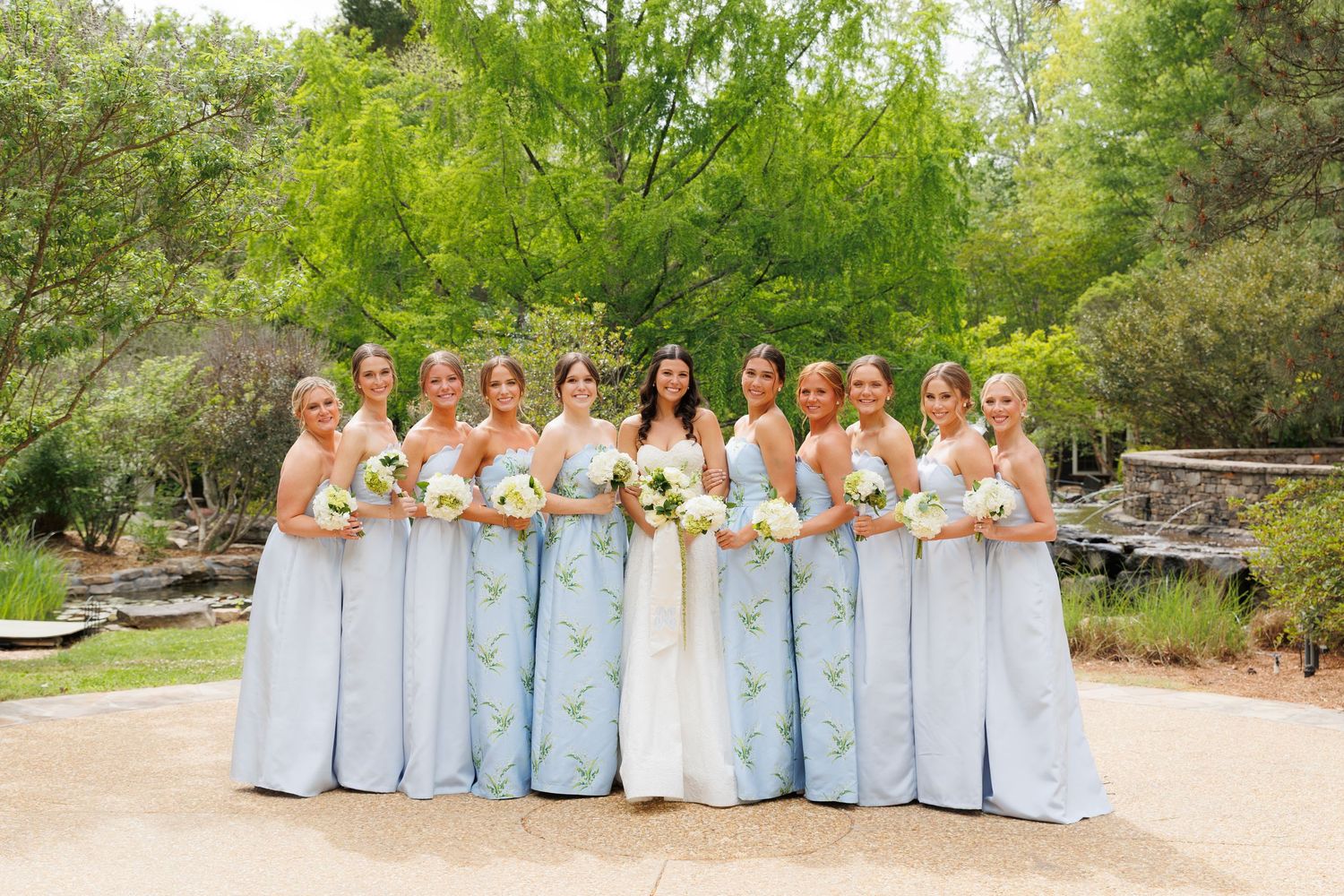 Bride and bridesmaids in light blue floral gowns holding white bouquets outdoors at a garden wedding venue.