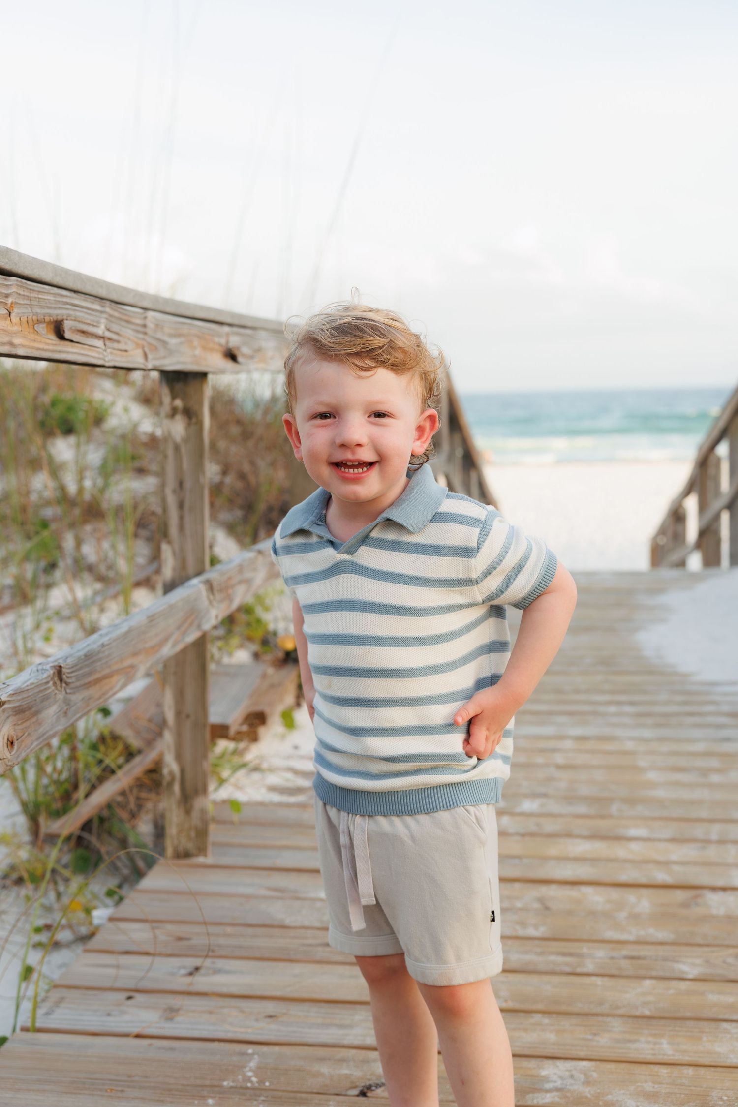 Smiling toddler boy in striped polo shirt stands on wooden beach boardwalk with ocean in background.