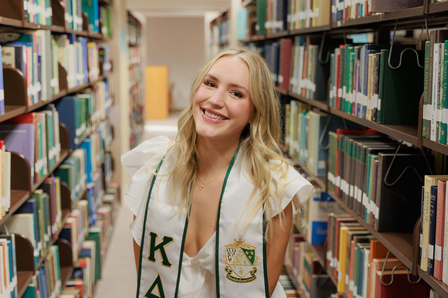 Smiling young woman wearing Kappa Delta sorority sash poses in college library aisle surrounded by bookshelves.