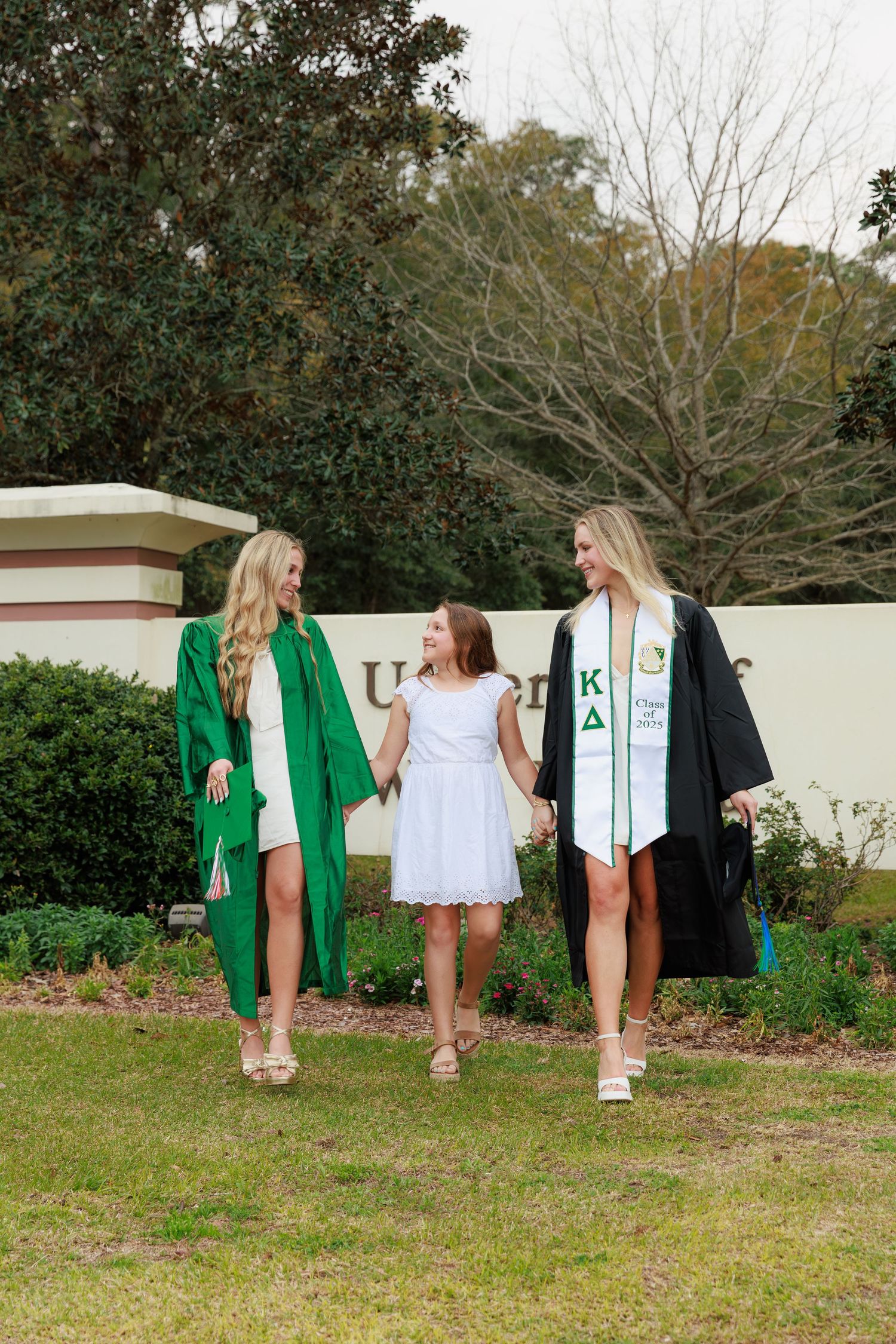 Two graduates in green and black robes walk with a young girl near a university sign on graduation day.