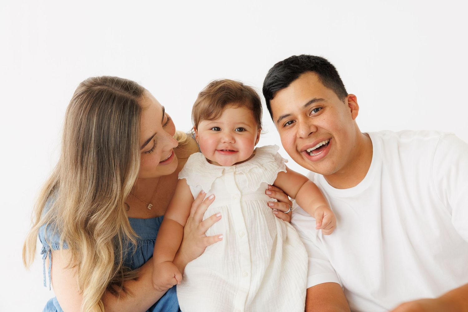 A happy young family with a smiling baby girl in white dress poses together against a white background.