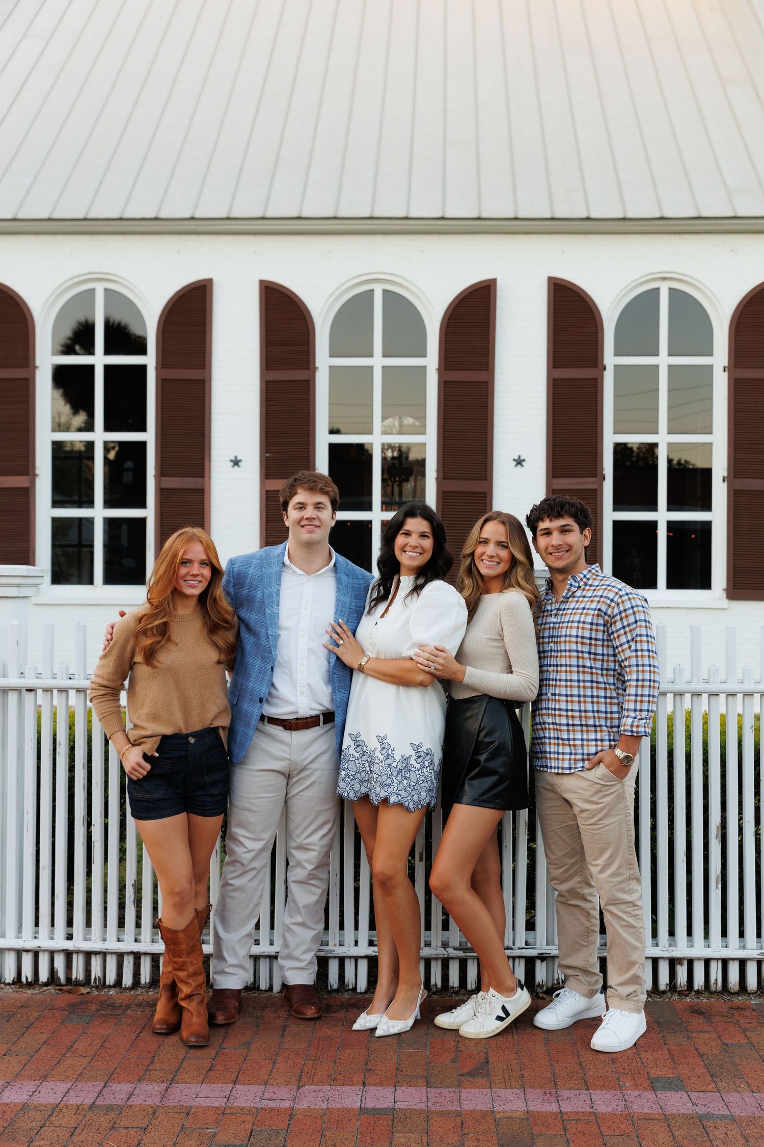 Group of five young adults smiling together outdoors by a white picket fence and historic building.