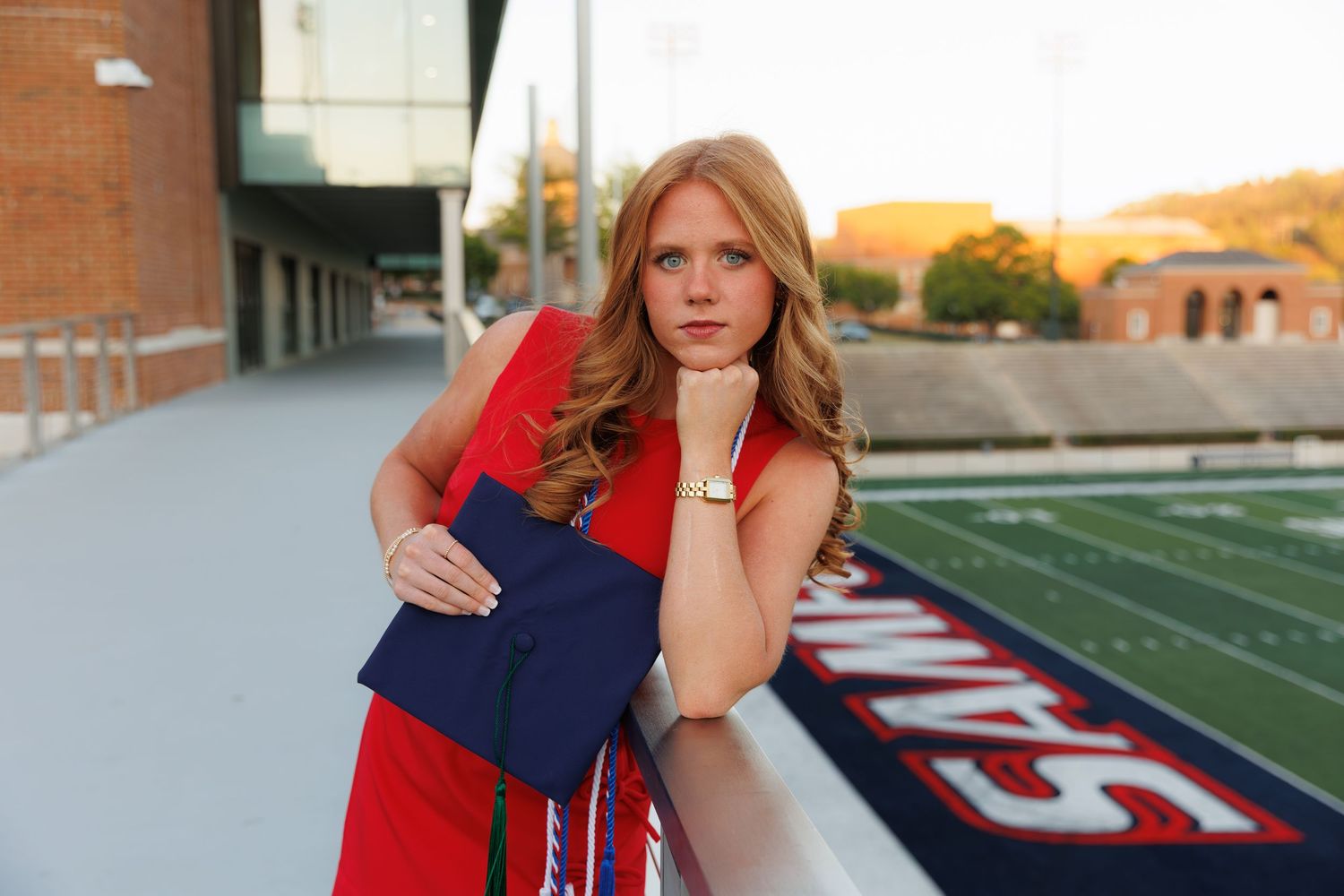 Smiling graduate in red dress holds navy cap while leaning on stadium railing at sunset.