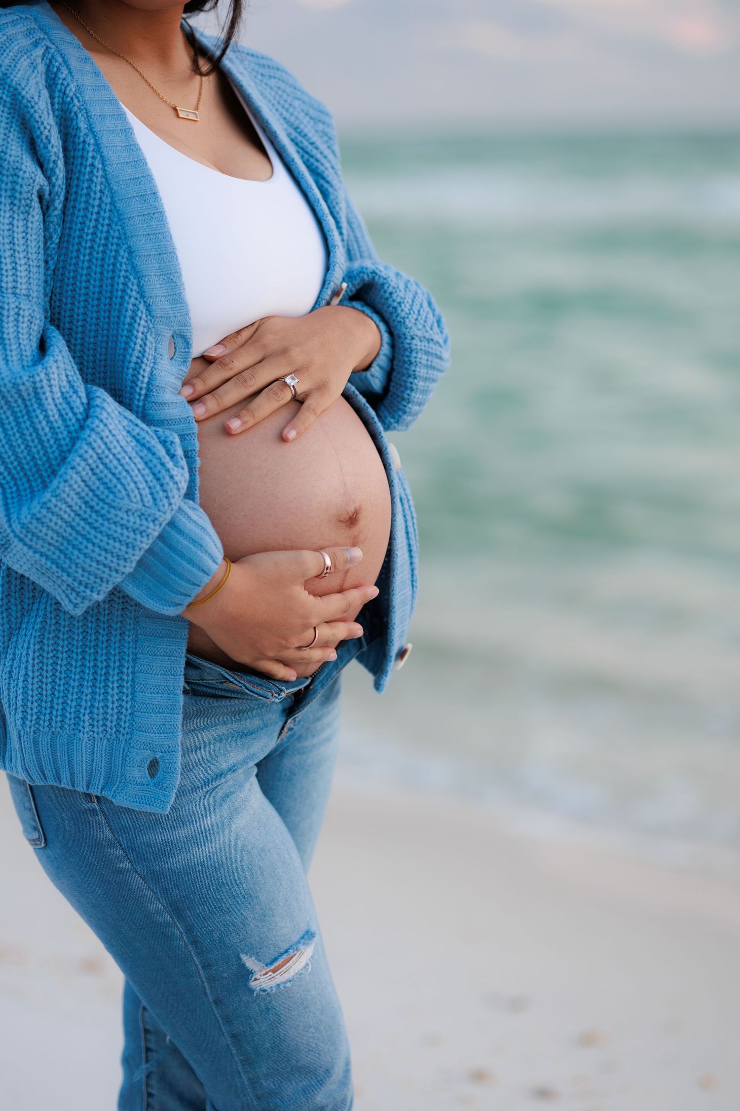 Close up of pregnant belly with hands cradling bump, wearing blue sweater and distressed jeans by ocean.