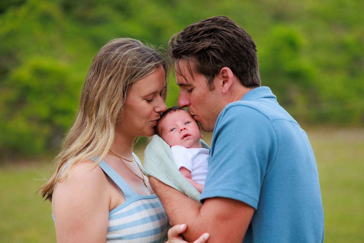 A couple shares a tender moment with their newborn baby outdoors against a green natural background.