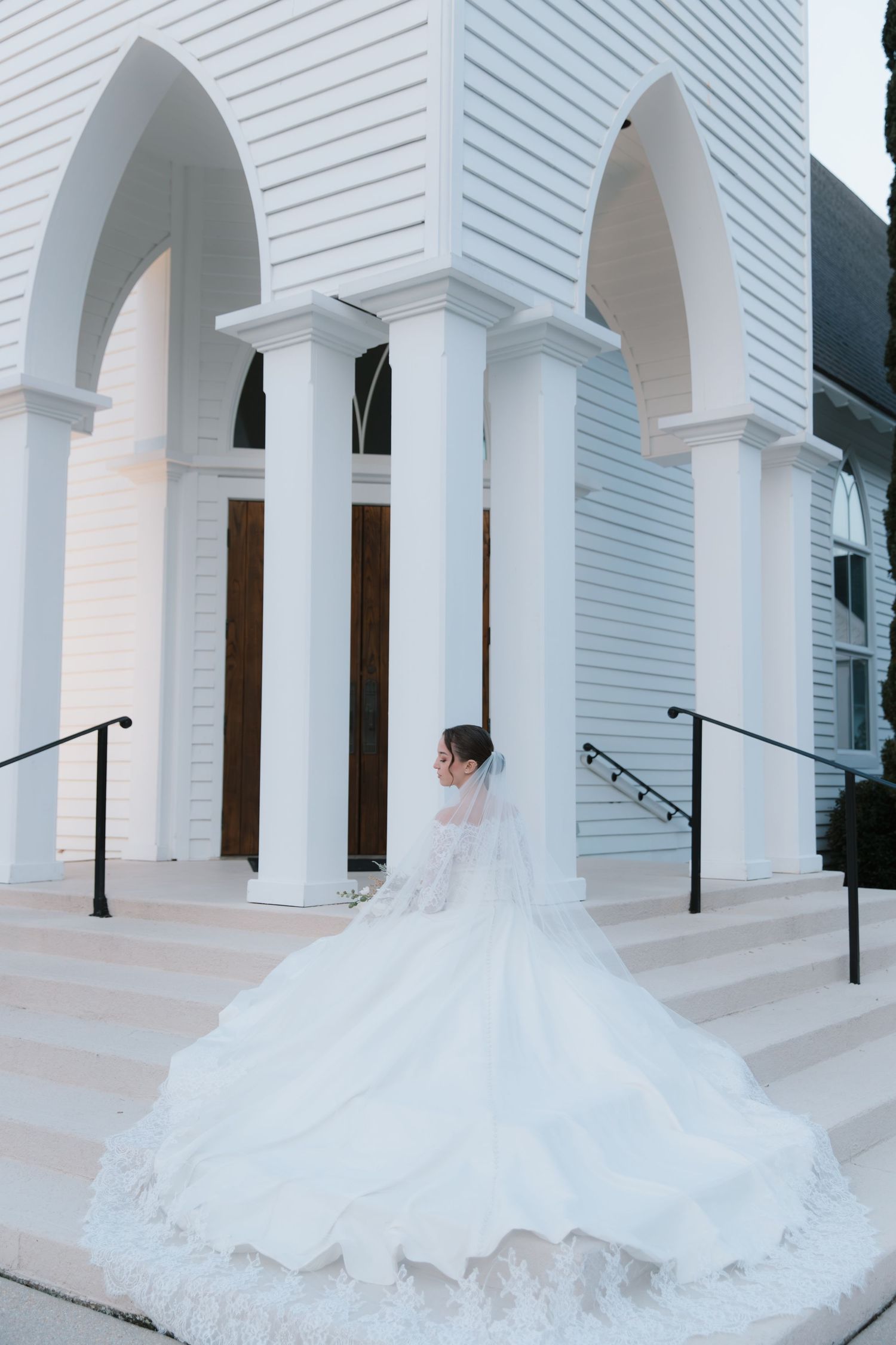 A flowing white wedding gown with a long train cascades down the steps of a white church with elegant arched entrances.