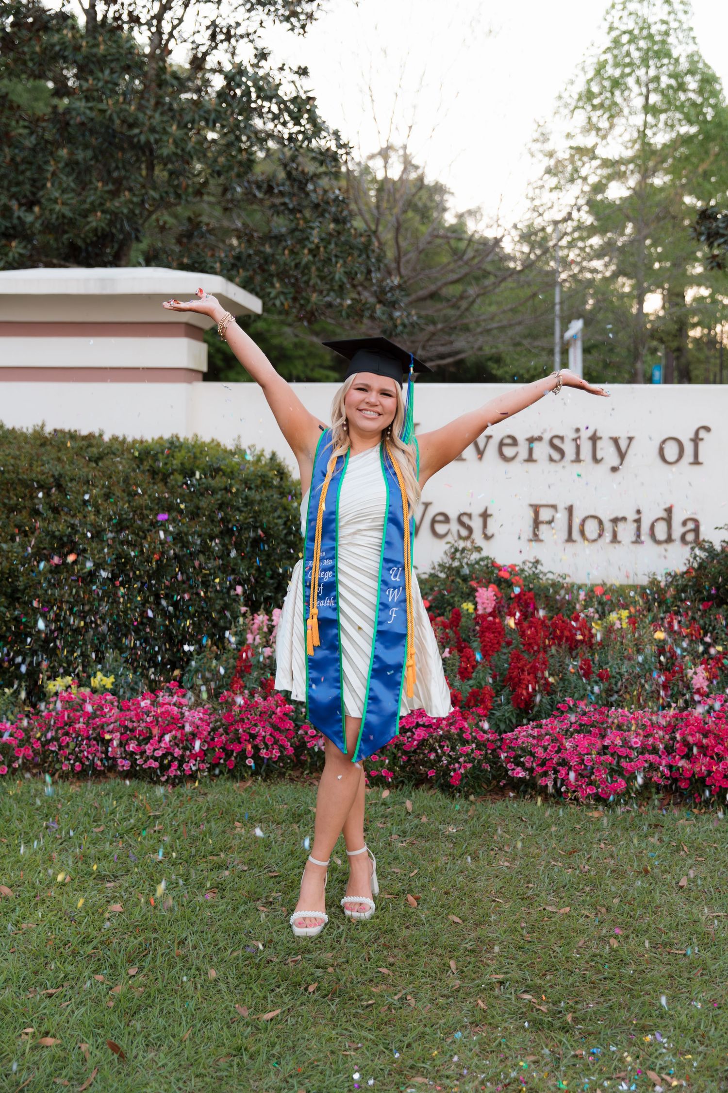 Graduate in cap and gown celebrates near University of West Florida sign with pink flowers.