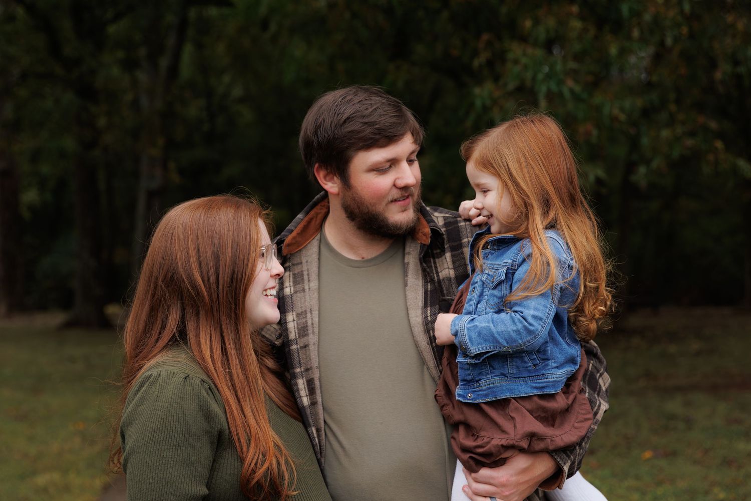 A family in casual clothing stands together outdoors against a wooded background during autumn.