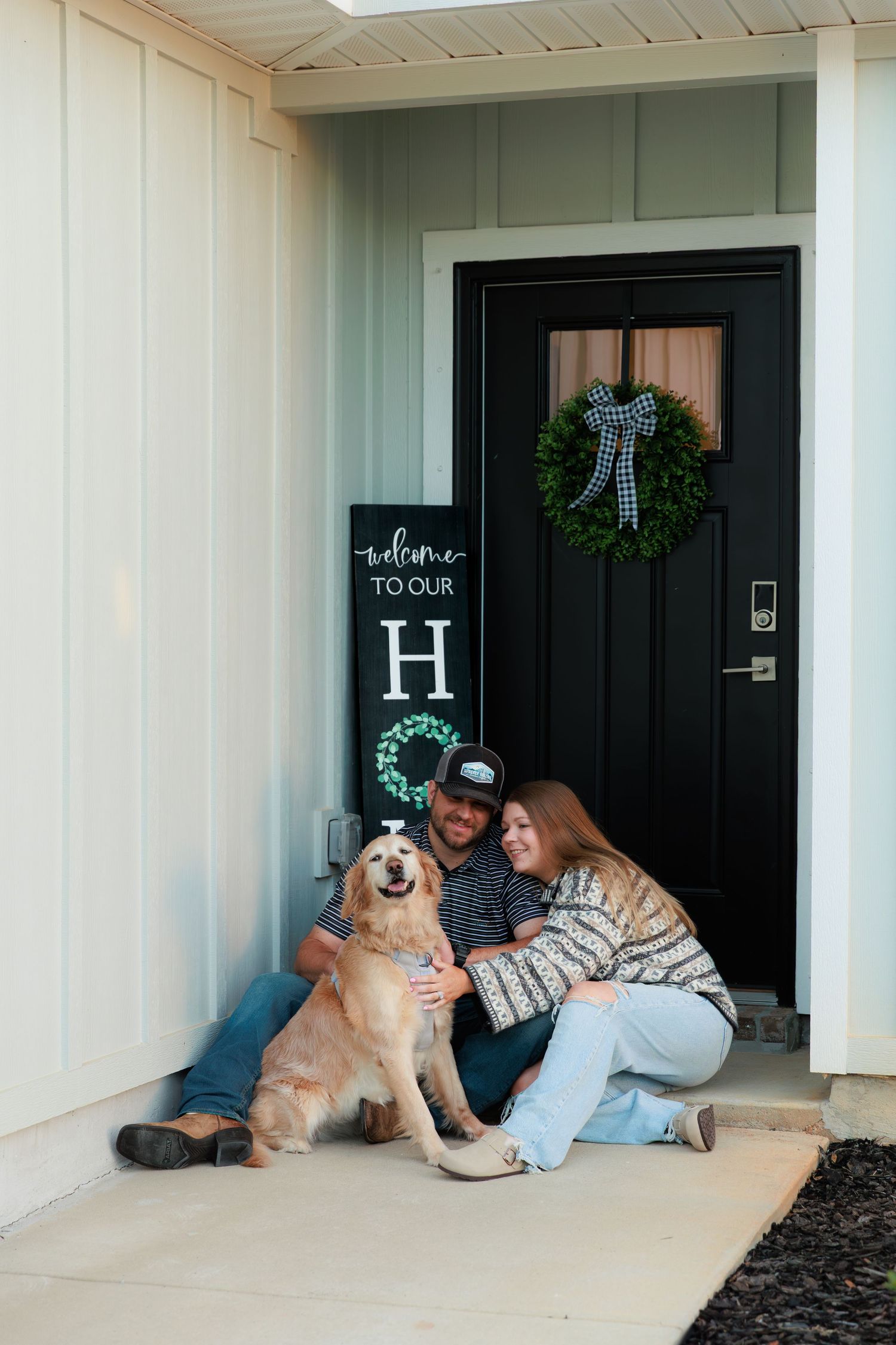 A golden retriever sits with someone on a front porch decorated with a wreath and welcome sign.