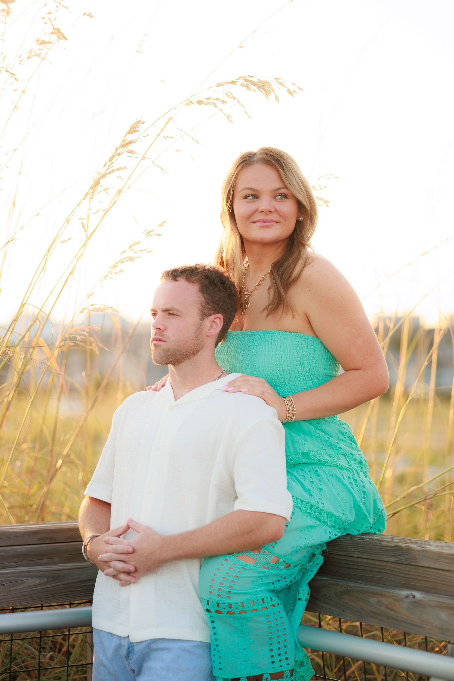 A romantic couple embraces on a wooden bench at sunset with tall beach grass in the background.