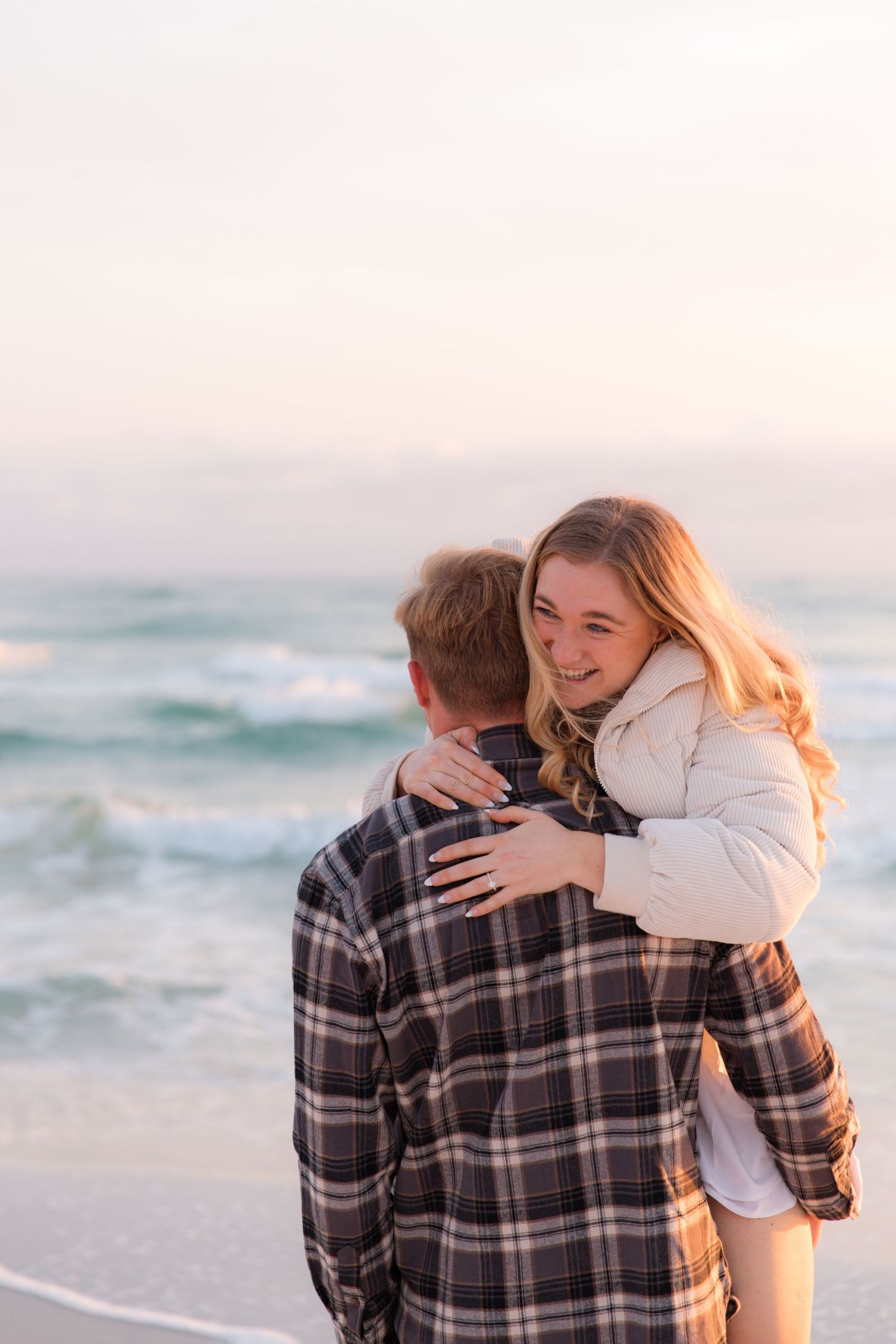 A couple embraces on a beach at sunset with waves crashing in the background.