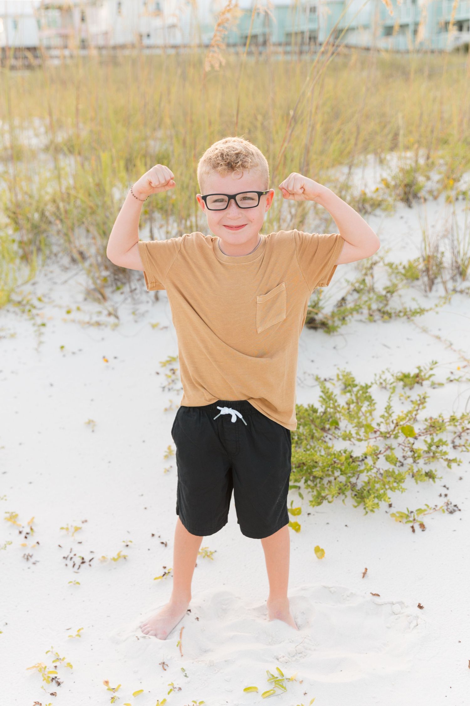Child in tan shirt and black shorts flexes muscles playfully on sandy beach.