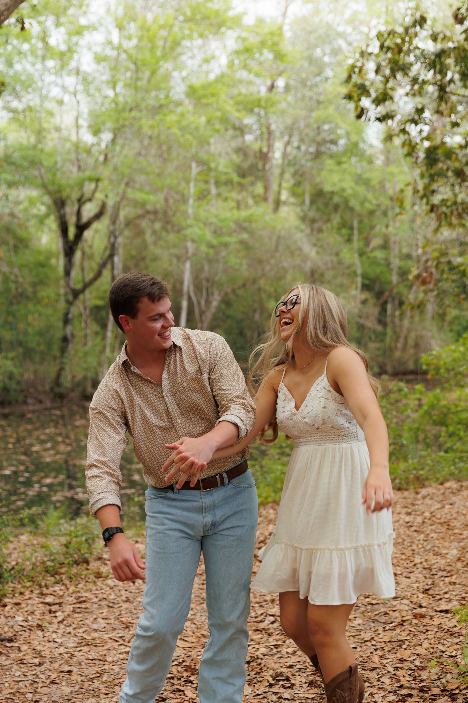 A laughing couple walks hand-in-hand through a wooded path, wearing casual country-style outfits.