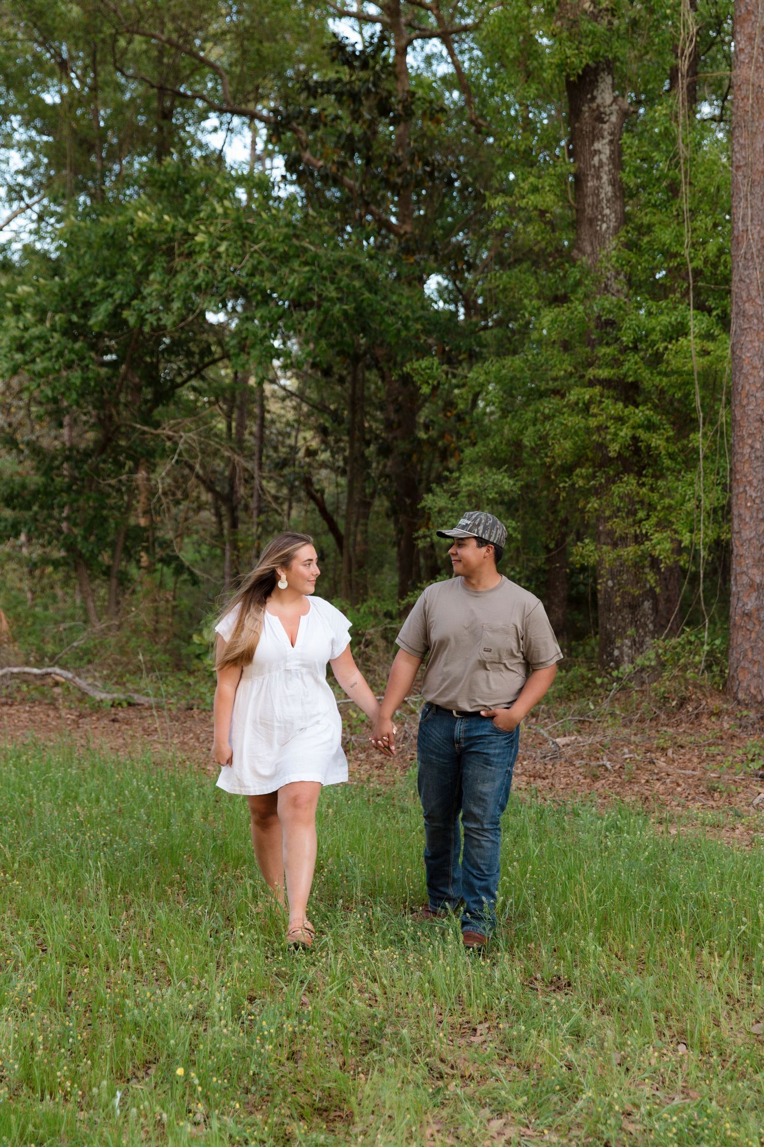 Couple holding hands walking in a green forest, woman in white dress, man in camo hat and jeans.