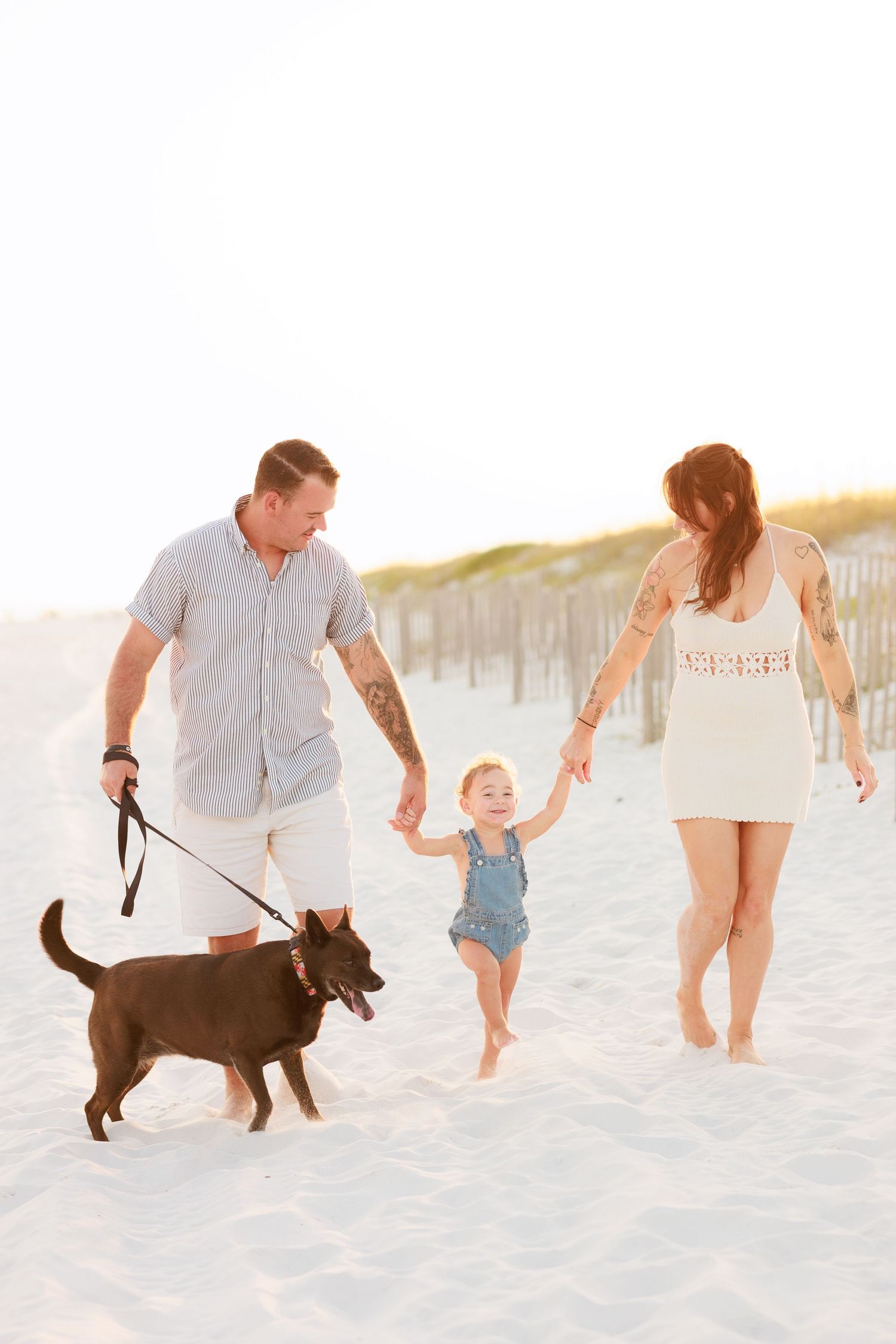 Happy family walking on white sand beach with toddler holding parents hands and dog on leash.
