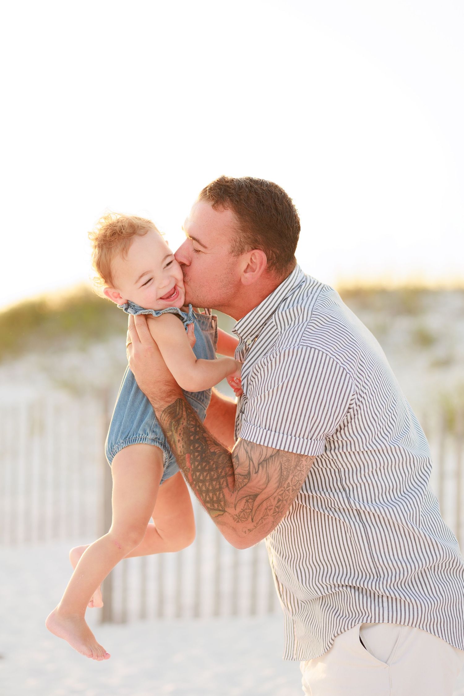A tattooed father kisses his laughing toddler on the cheek at a sunny beach with white sand dunes.