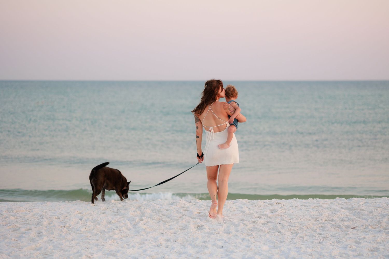 Woman in white dress holding baby while walking dark dog on leash along sandy beach at sunset.
