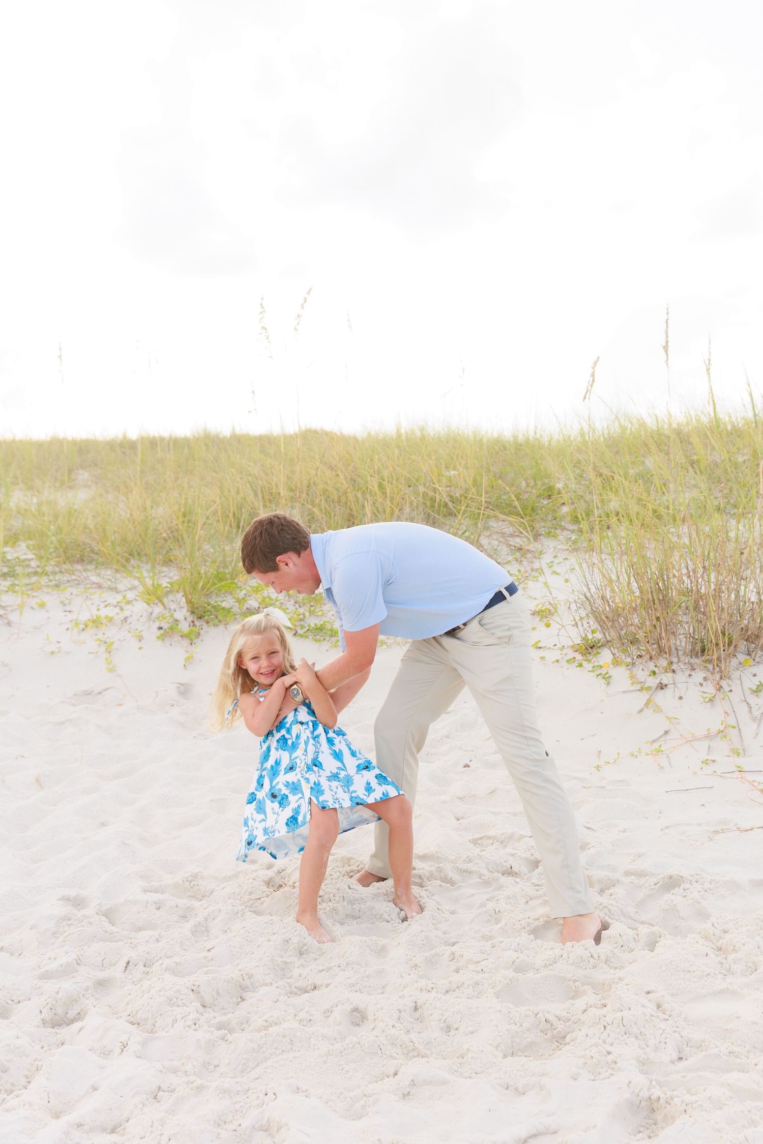 A father bends down to hug his smiling young daughter in a blue floral dress on a sunny beach.