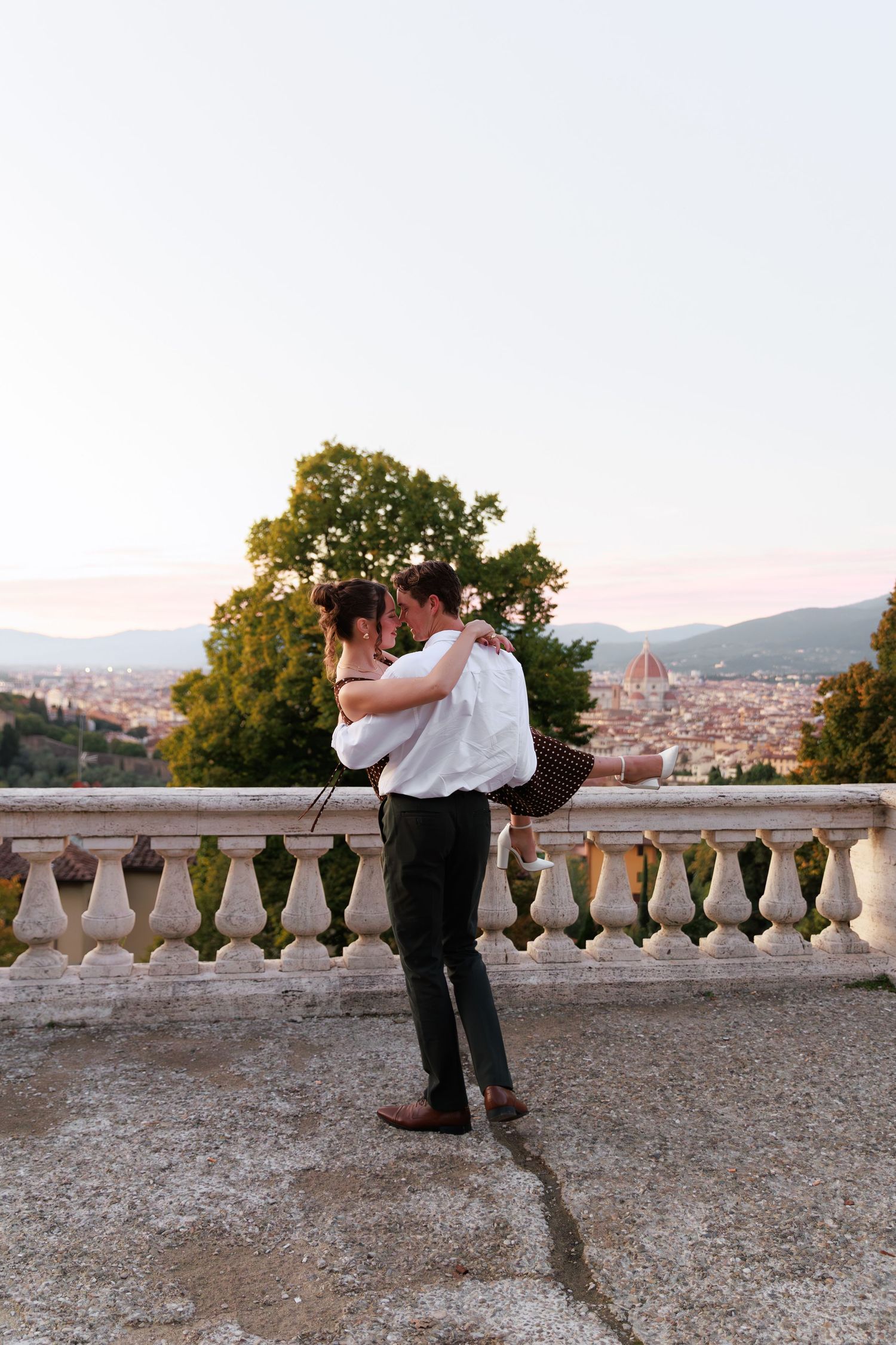 Couple sharing passionate kiss as man lifts woman on terrace overlooking Florence at dusk.
