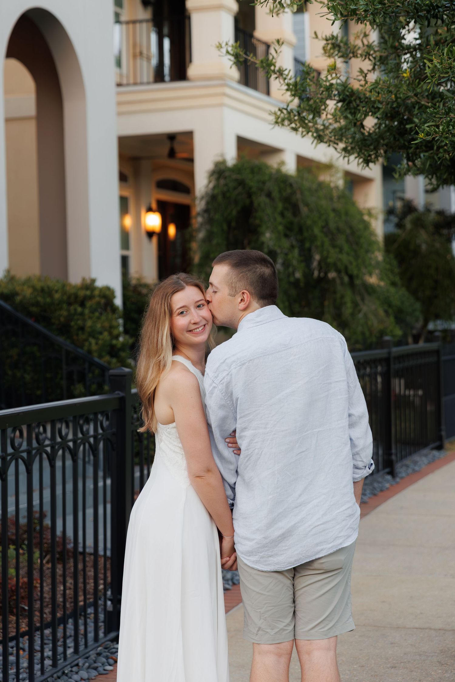 A couple shares a romantic kiss outdoors, woman in white dress smiling, man in light blue shirt, near elegant building.