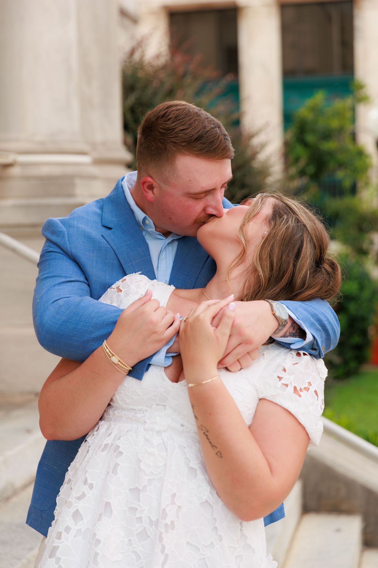 A couple shares a romantic kiss, the groom in a blue suit embracing the bride in a white lace dress outdoors.