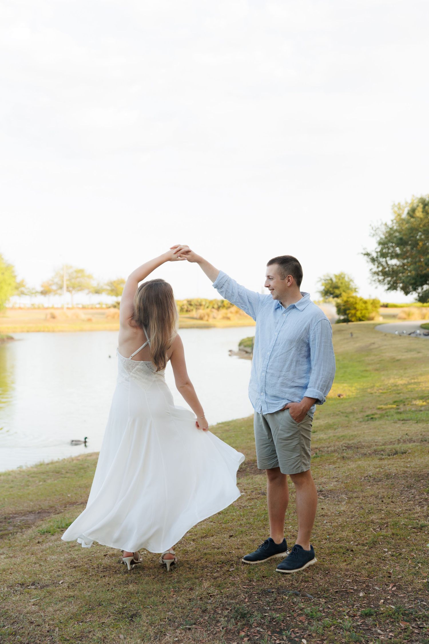 Couple dancing by a lakeside, woman in white dress being twirled by man in blue shirt and shorts.