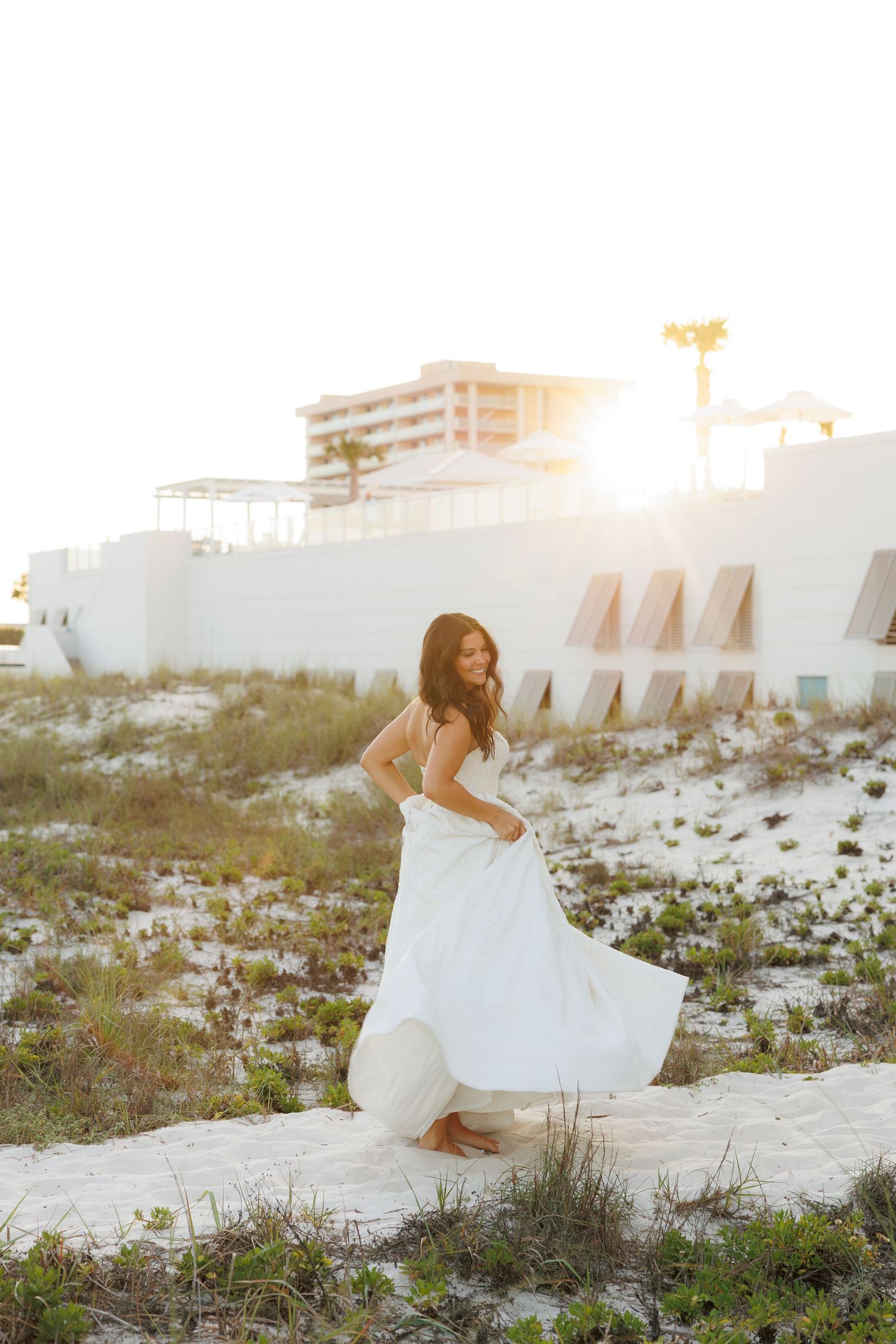 Bride in flowing white gown twirling on white sand beach at golden sunset with coastal building backdrop.