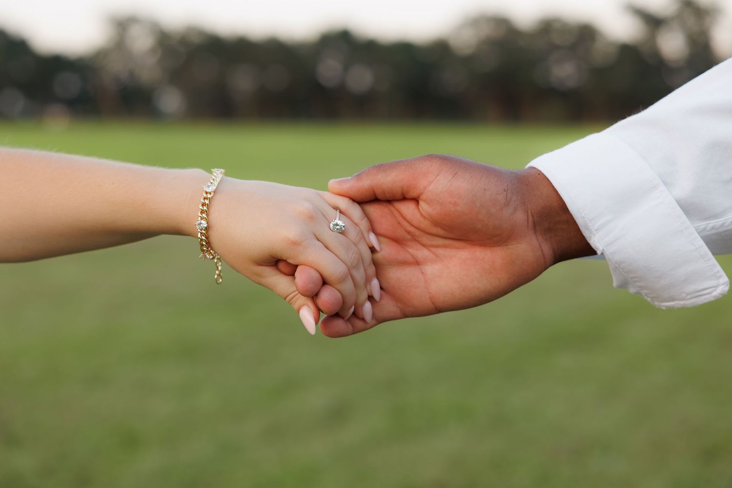 Close-up of interracial couple holding hands, woman wearing gold bracelet and engagement ring outdoors.