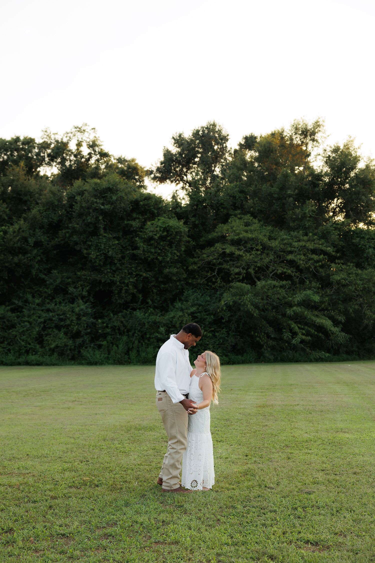 Couple sharing a romantic moment in a sunlit green field with lush trees in the background.