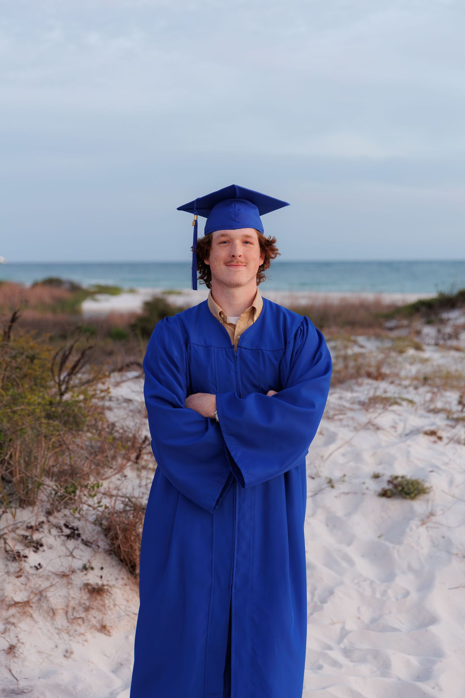 Young male graduate in blue cap and gown standing on beach with arms crossed.