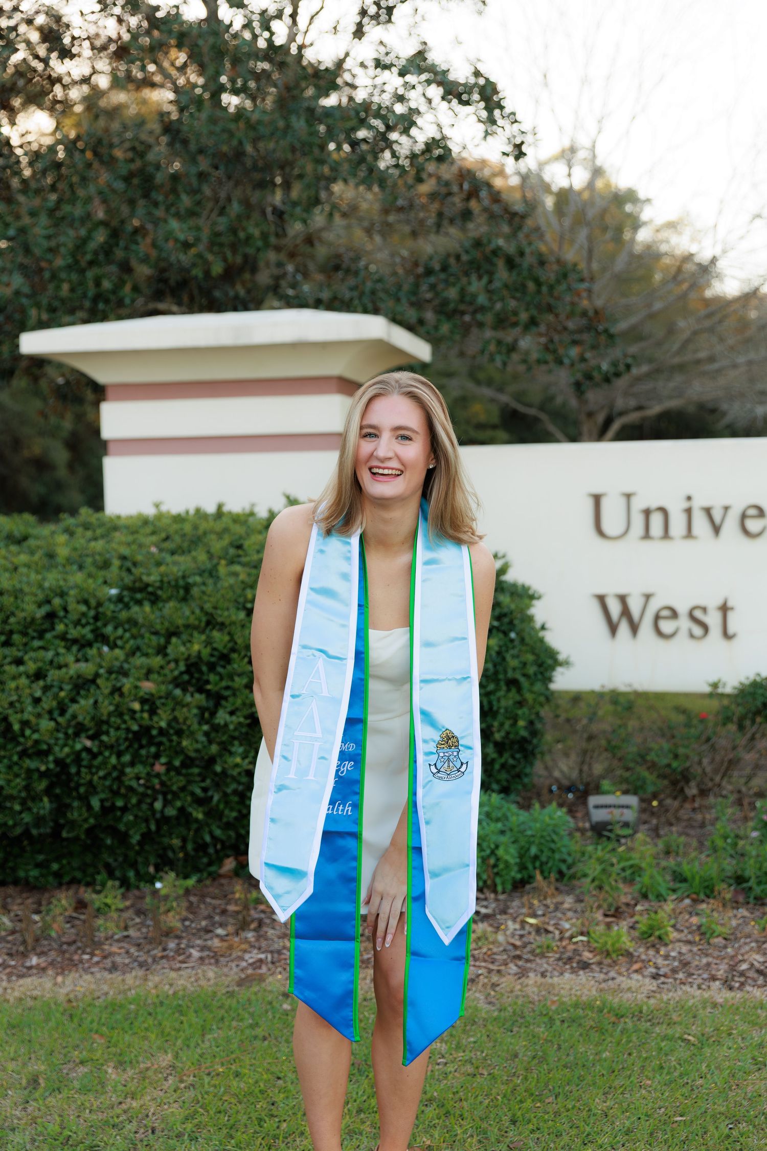 Graduate in light blue stoles and teal dress poses in front of university signage during commencement photo session.