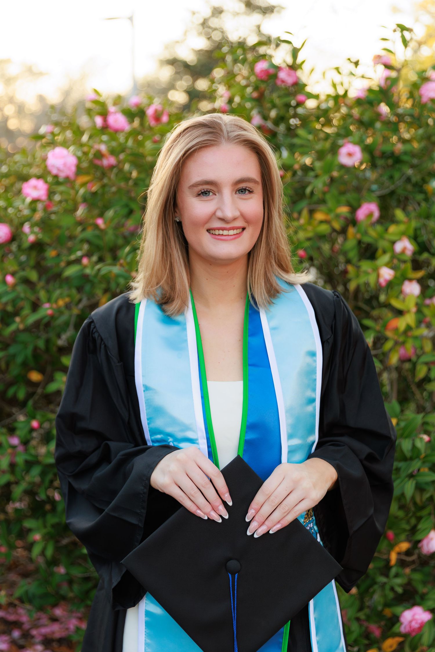 Graduate in black cap and gown with light blue stole poses in front of blooming rose bushes during graduation photos.