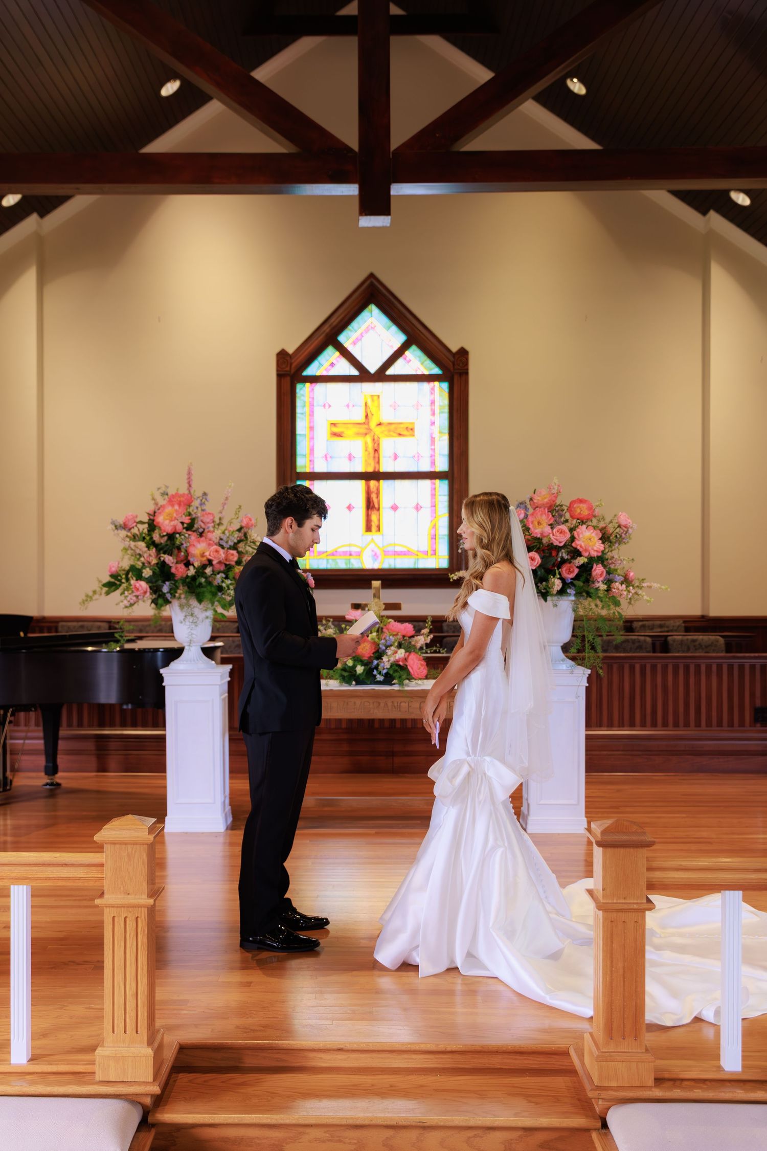 Two people stand together at the altar of a chapel with stained glass windows and pink floral arrangements.