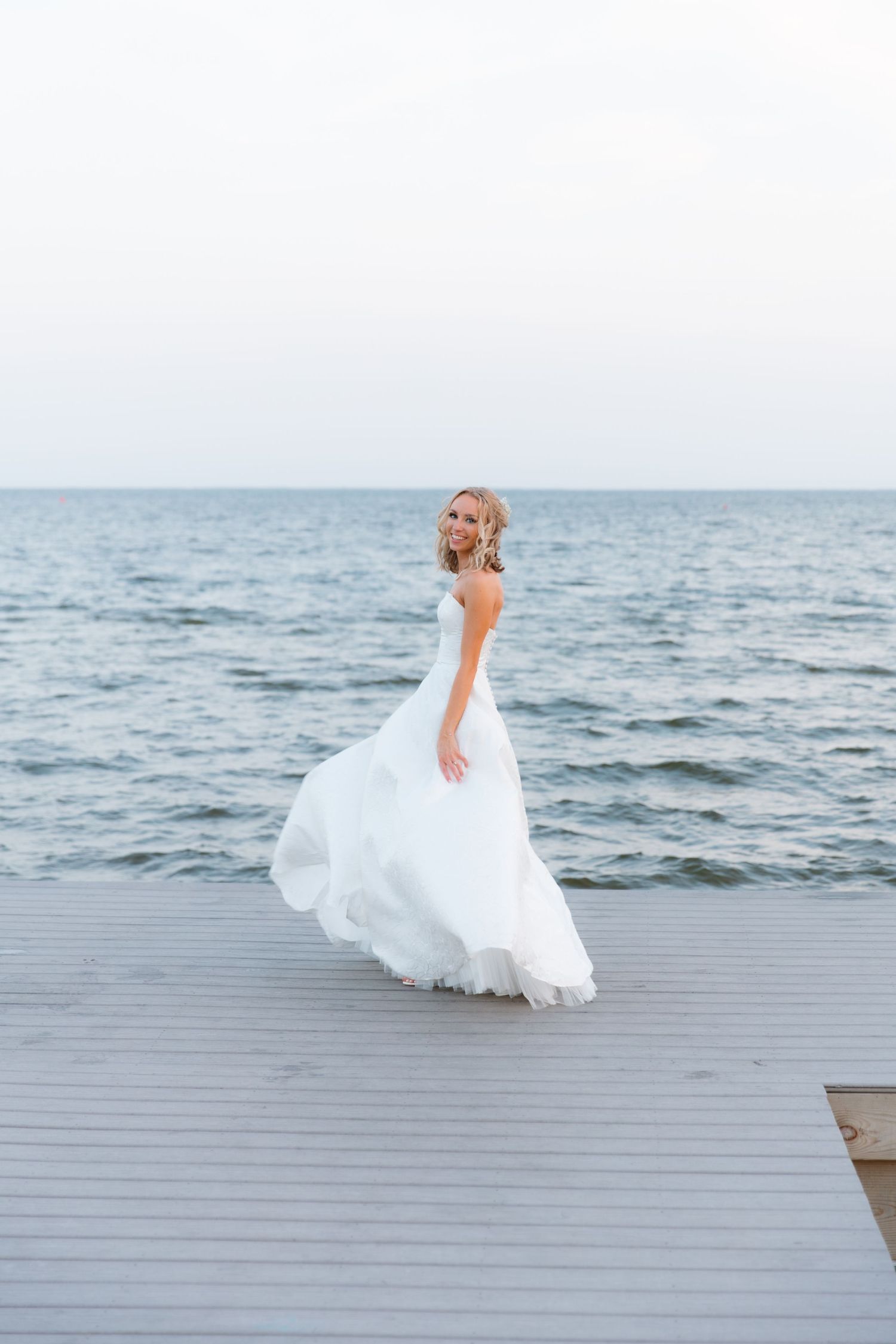 Flowing white wedding dress twirls on wooden dock by the ocean.