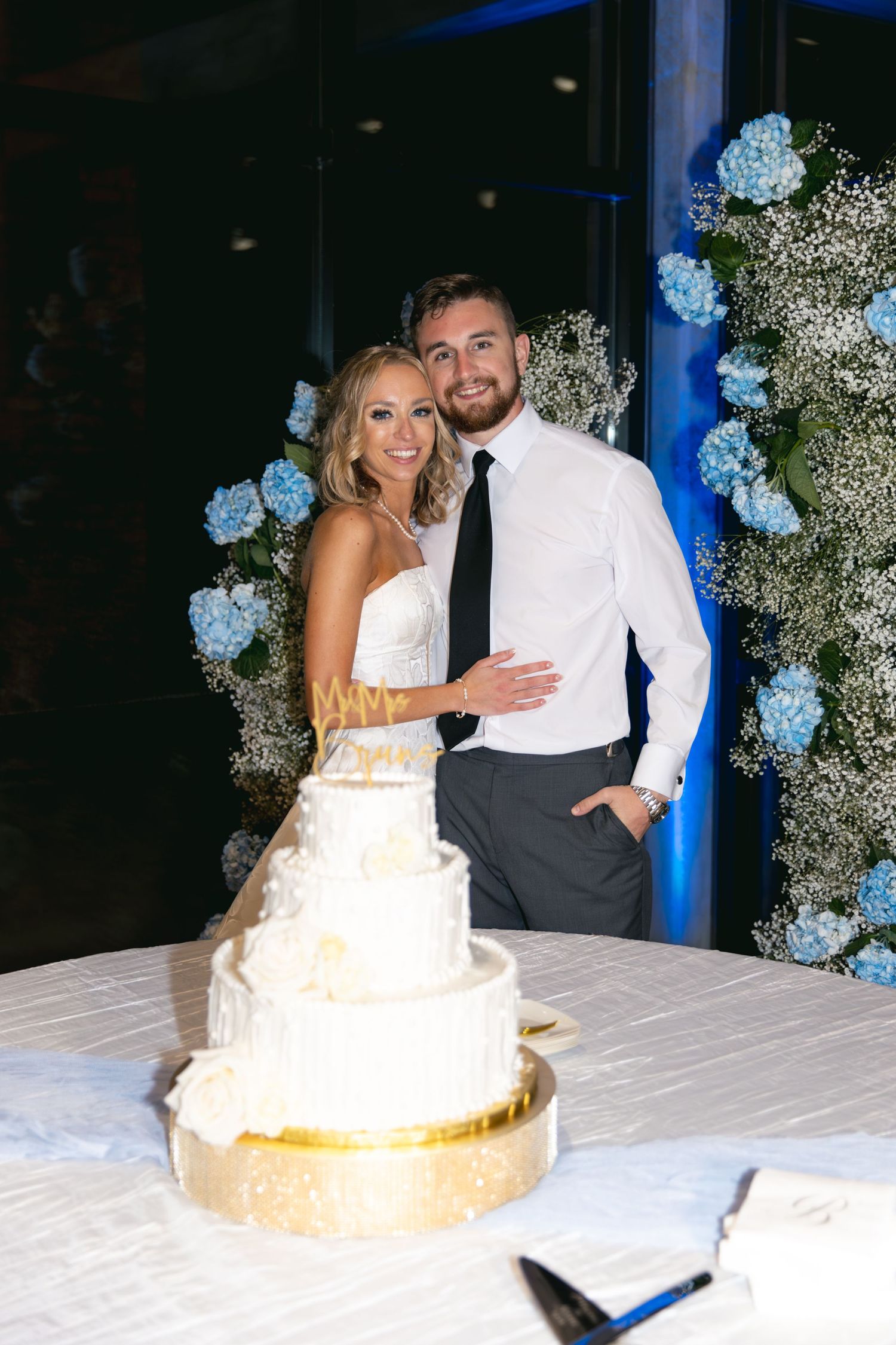 Happy couple poses behind their white wedding cake with blue flower backdrop.