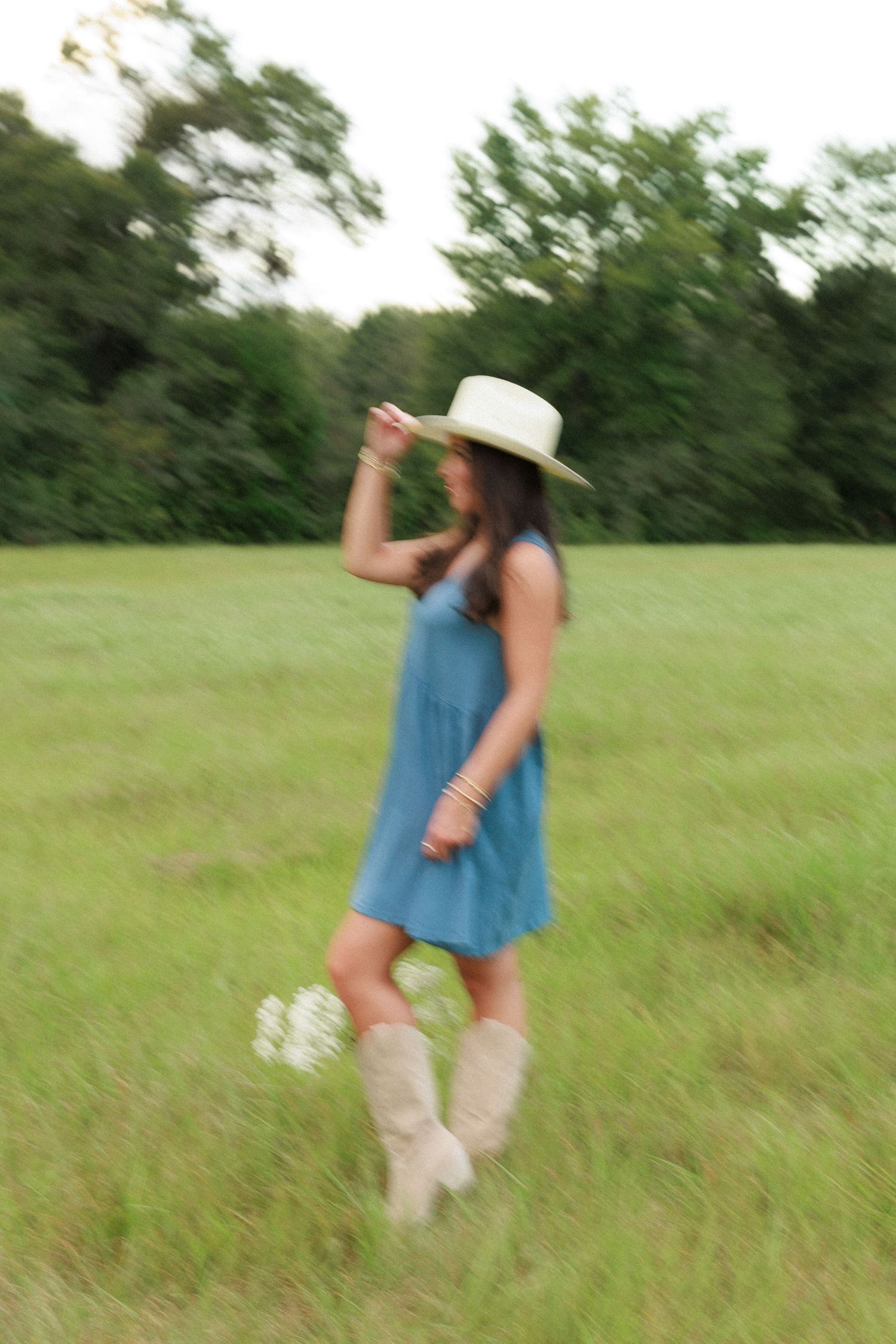 A dreamy summer portrait in a blue dress and cowboy boots posing playfully in a grassy field.