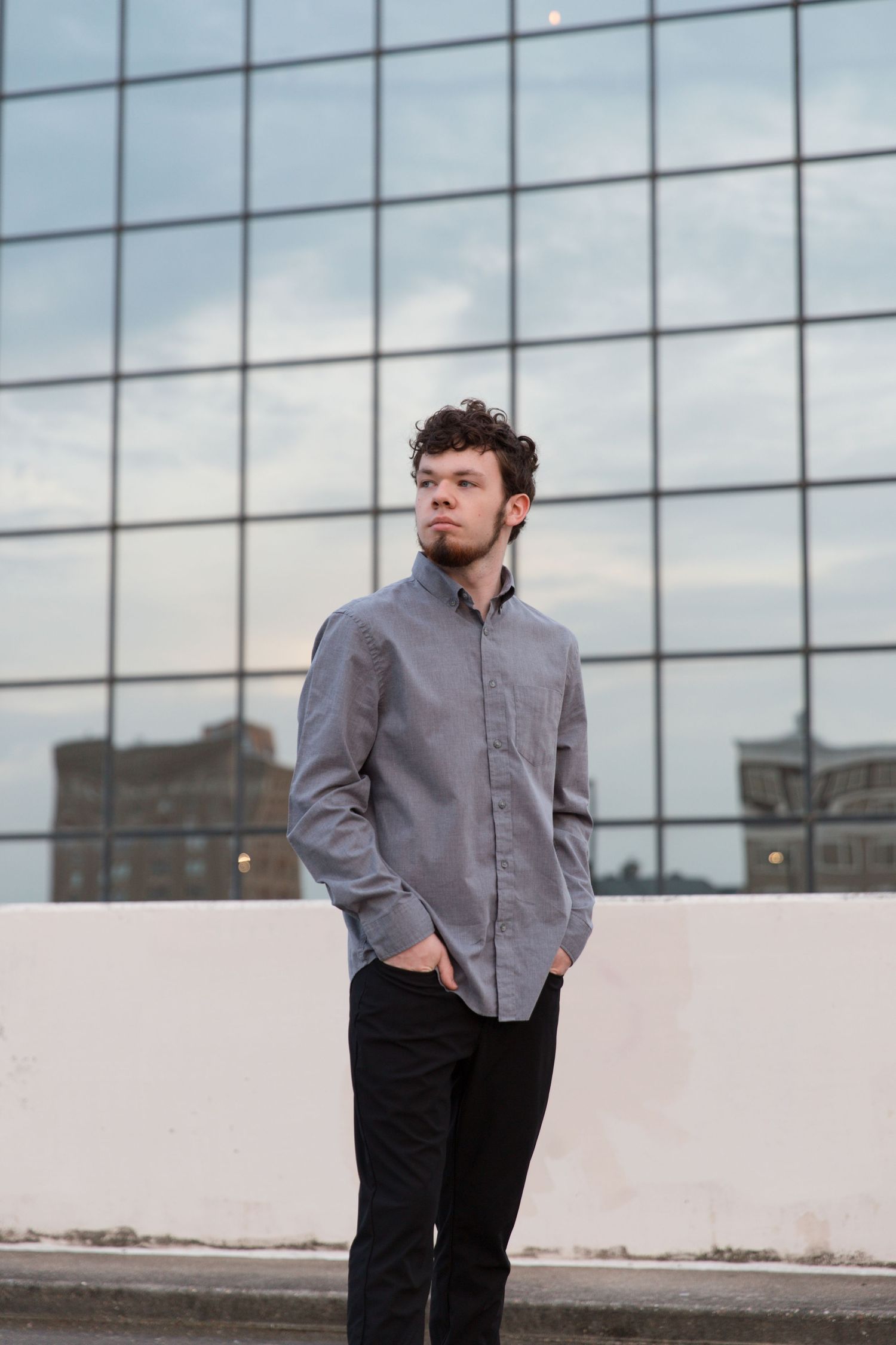 Professional portrait against modern glass building facade wearing grey button down shirt and black pants.