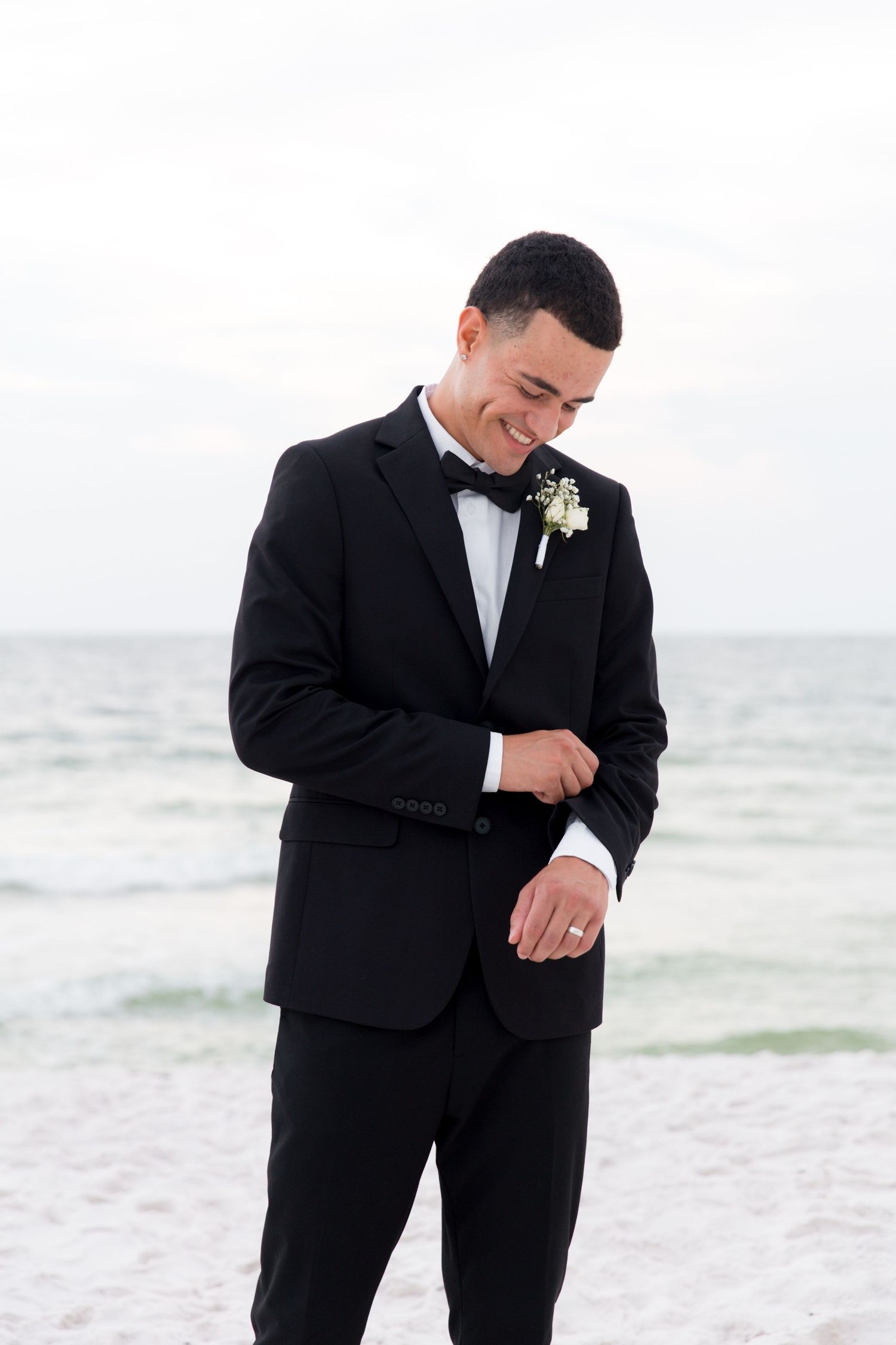 Groom in classic black tuxedo smiling and adjusting sleeve while standing on beach at sunset.